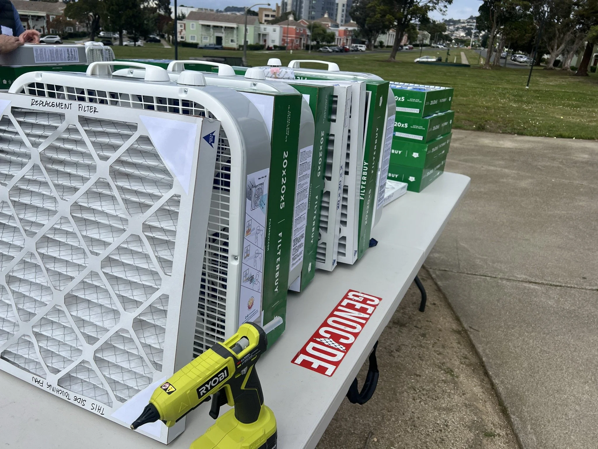 A folding table is on a sidewalk with green space in the background. The table has a red sticker that reads "Genocide" with the "I" in the shape of Palestine. On top of the table is a hot glue gun, as well as multiple DIY air purifiers and filters.