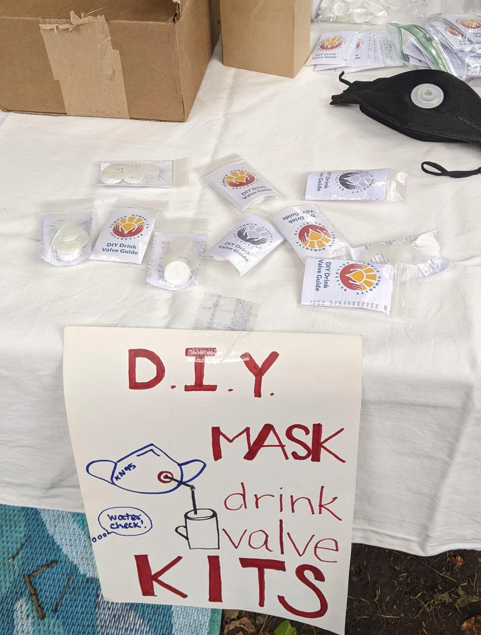 A table with a hand-drawn sign hanging over the front. The sign reads "D.I.Y. MASK drink valve KITS" and has an image of a KN95 mask. The KN95 has a drink valve installed and a straw coming out of the valve and into a cup. A thought bubble on the sig