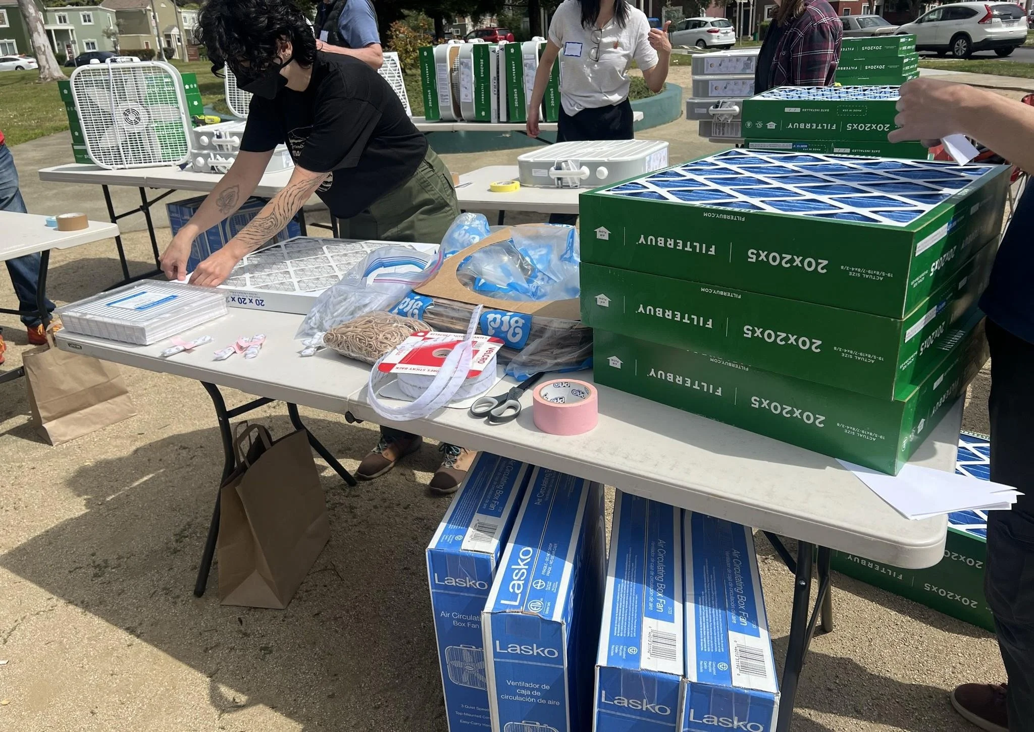 One masked person applying stickers to an air filter on a folding table. Beside the filter is multiple DIY air purifier materials, including cardboard shrouds, tape, and filters.