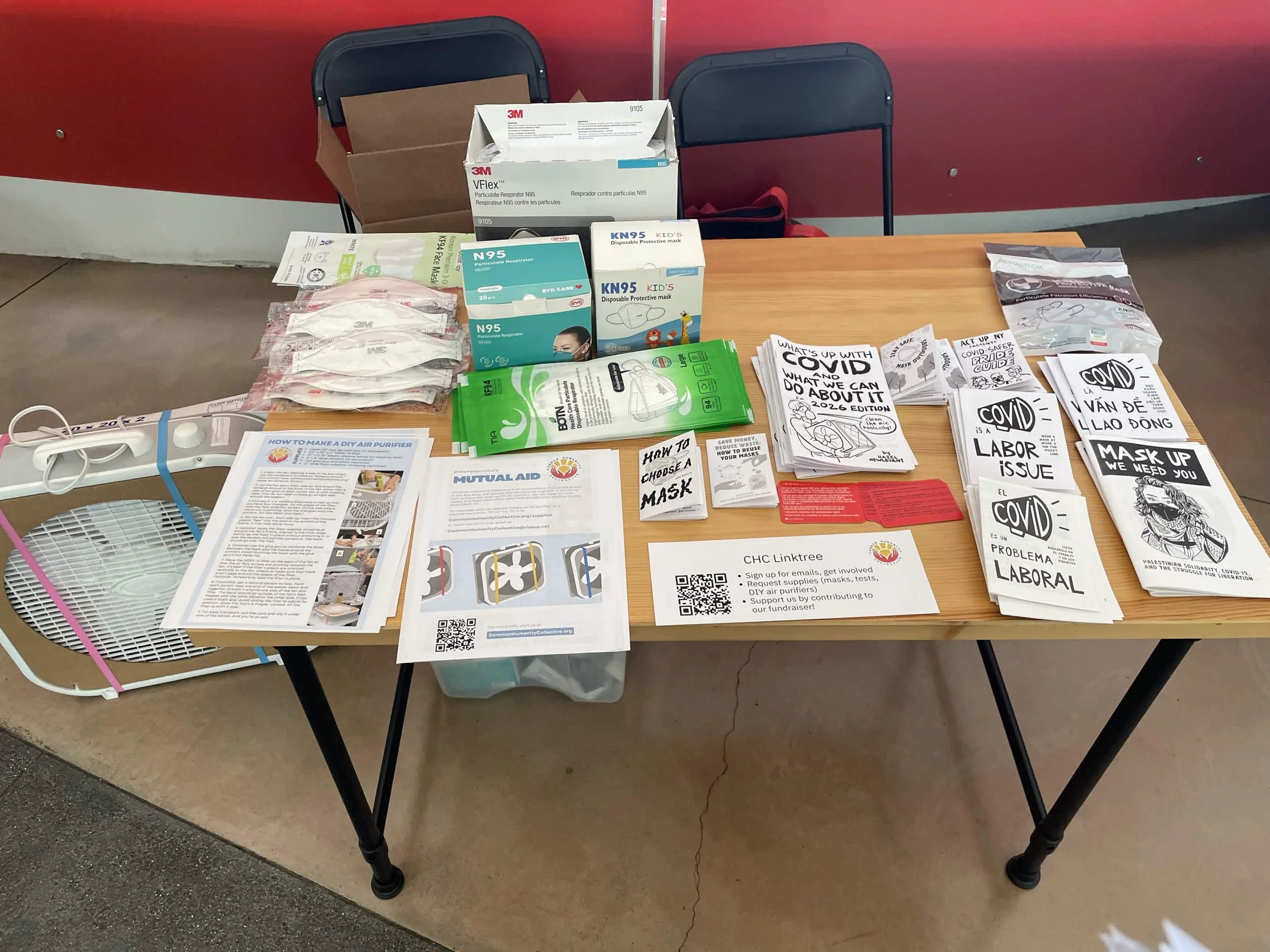 A rectangular table with numerous zines and masks covering it. There are two CHC flyers displayed, including a step-by-step guide to assembling a DIY air purifier. On the ground beside the table is a DIY air purifier. 