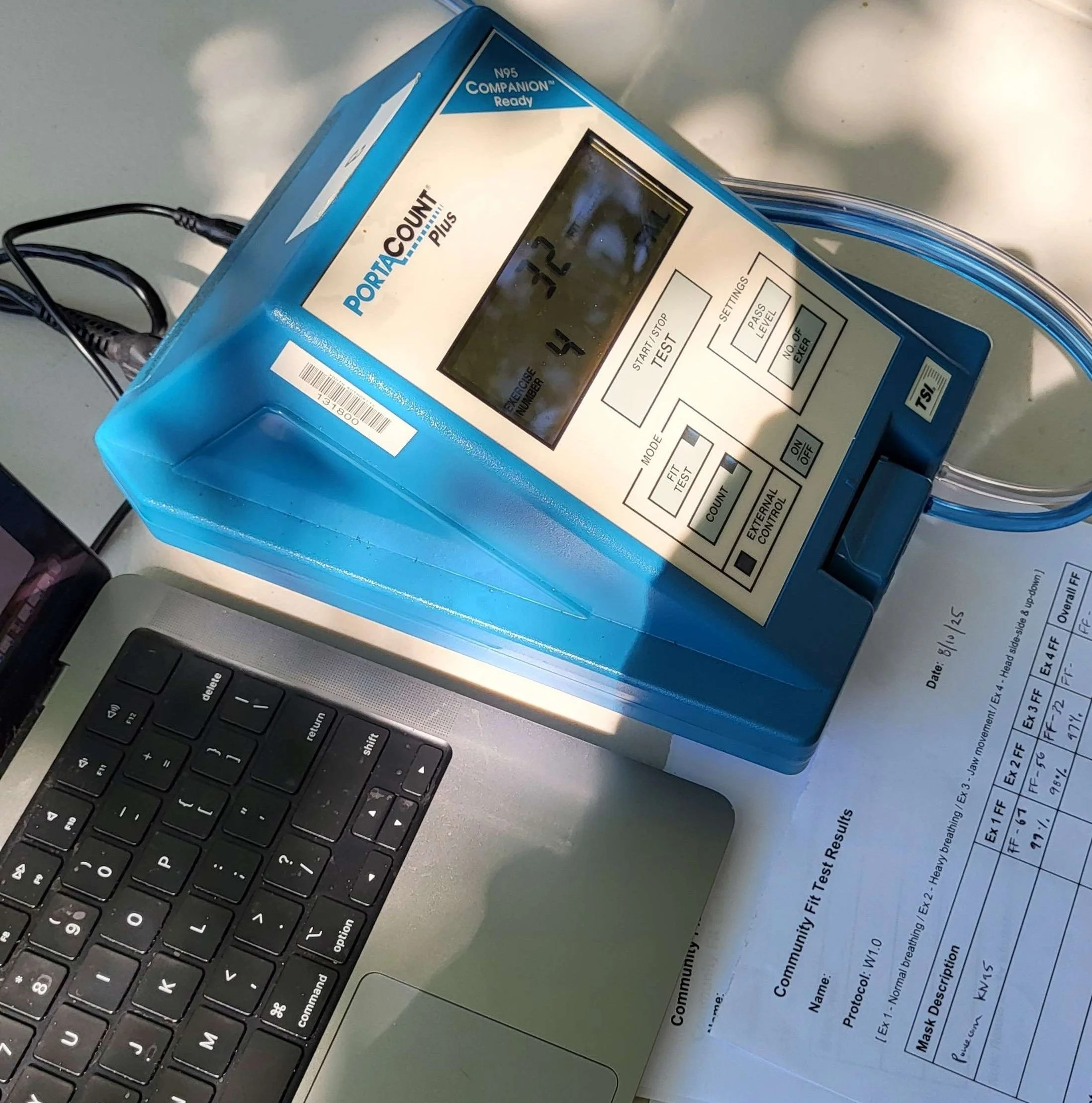 On a foldout table is a blue and white portacount plus machine, with the words "N95 Companion Ready" in the top righthand corner. To the side is an open laptop and a piece of paper that reads "Community Fit Test Results" partially filled out with dat
