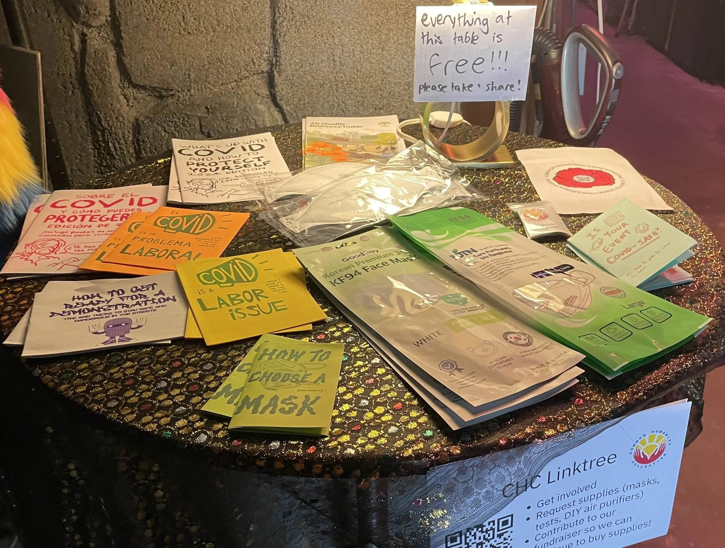 A circular table with numerous zines, masks, and a CHC DIY drink valve kit covering it. A lamp is on the table with a piece of paper taped to it that reads "everything on this table is free!! please take + share!" 