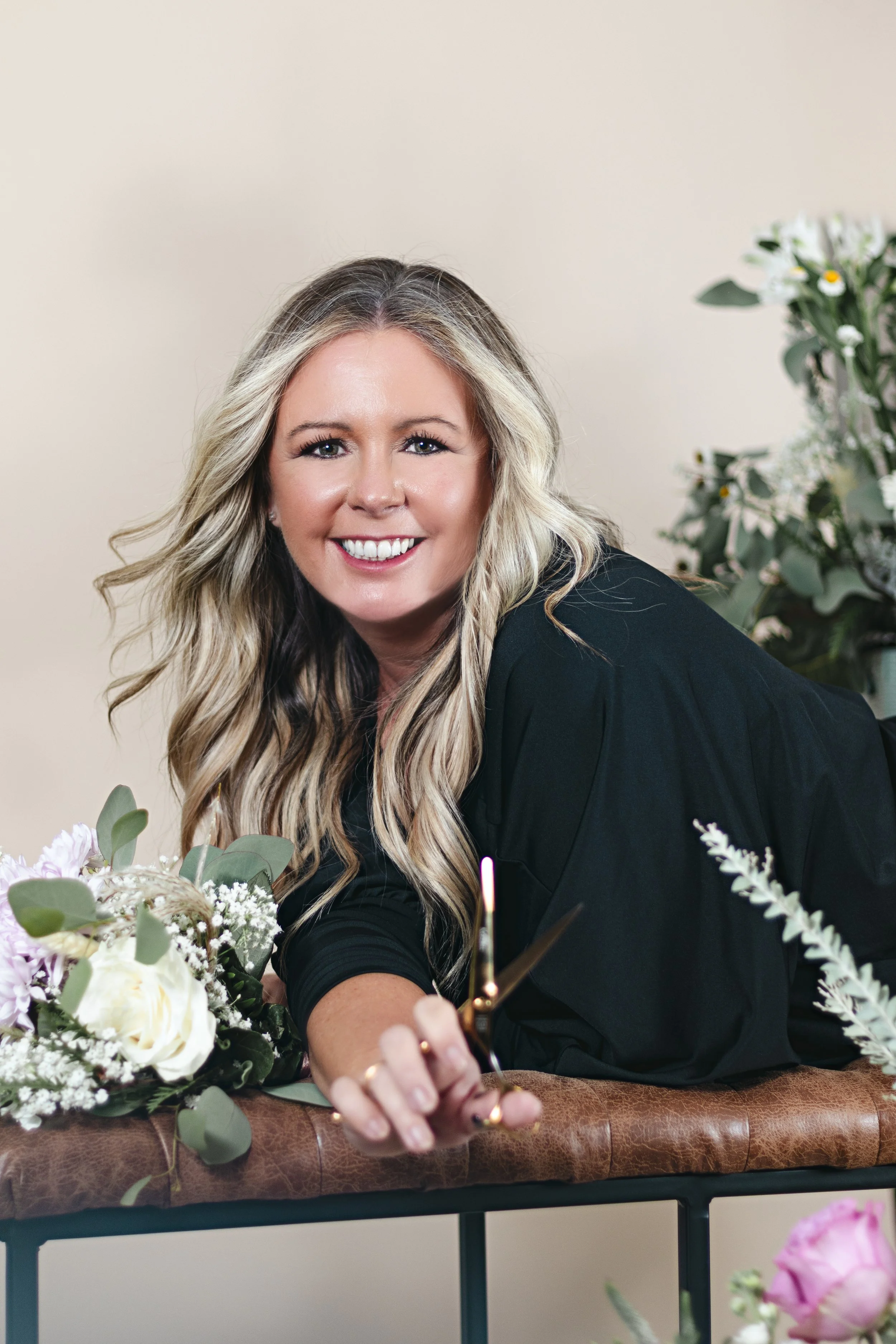 Woman posing laying on bench in front of white backdrop. The woman is blonde with softly curled hair and she is holding shears.