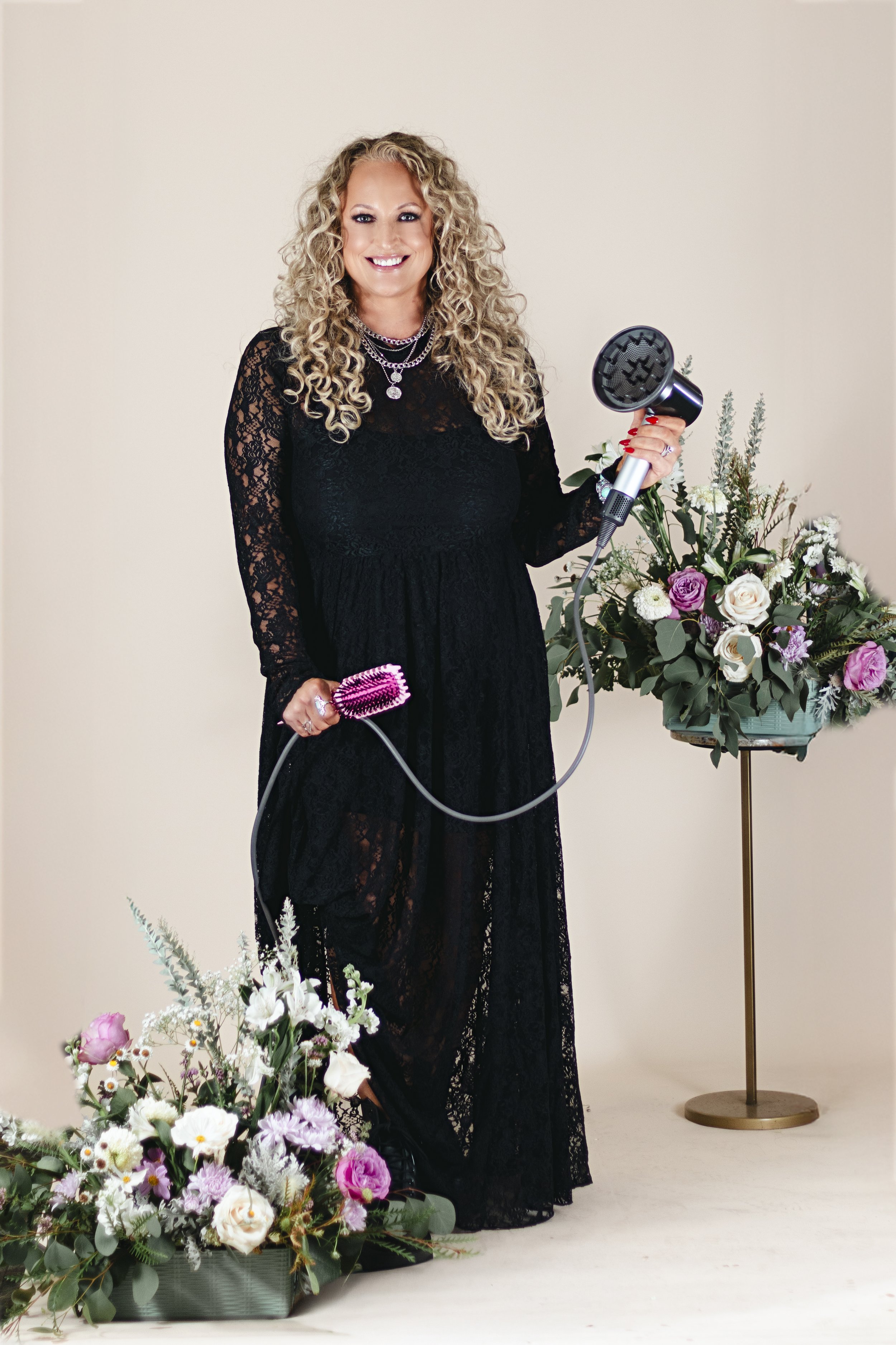 Woman posing infront of cream backdrop. The woman has curly blonde hair and is wearing a black lace dress. She is holding a hair brush and blow dryer. Flowers stand in the foreground and background.