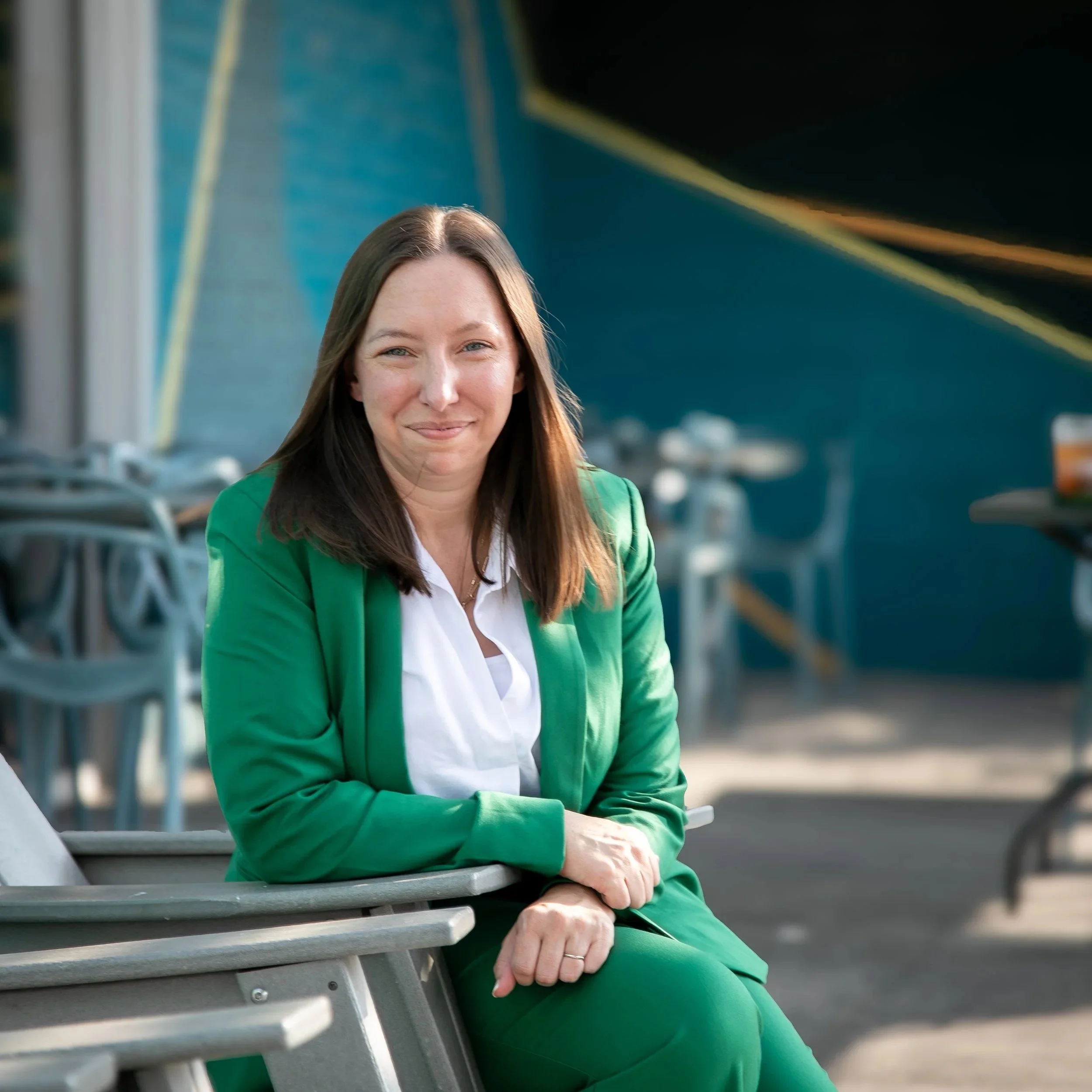 A woman with long brown hair sitting on a bench at an outdoor stadium or park, wearing a green jacket over a white shirt, smiling gently, with metal stadium seats visible in the background.