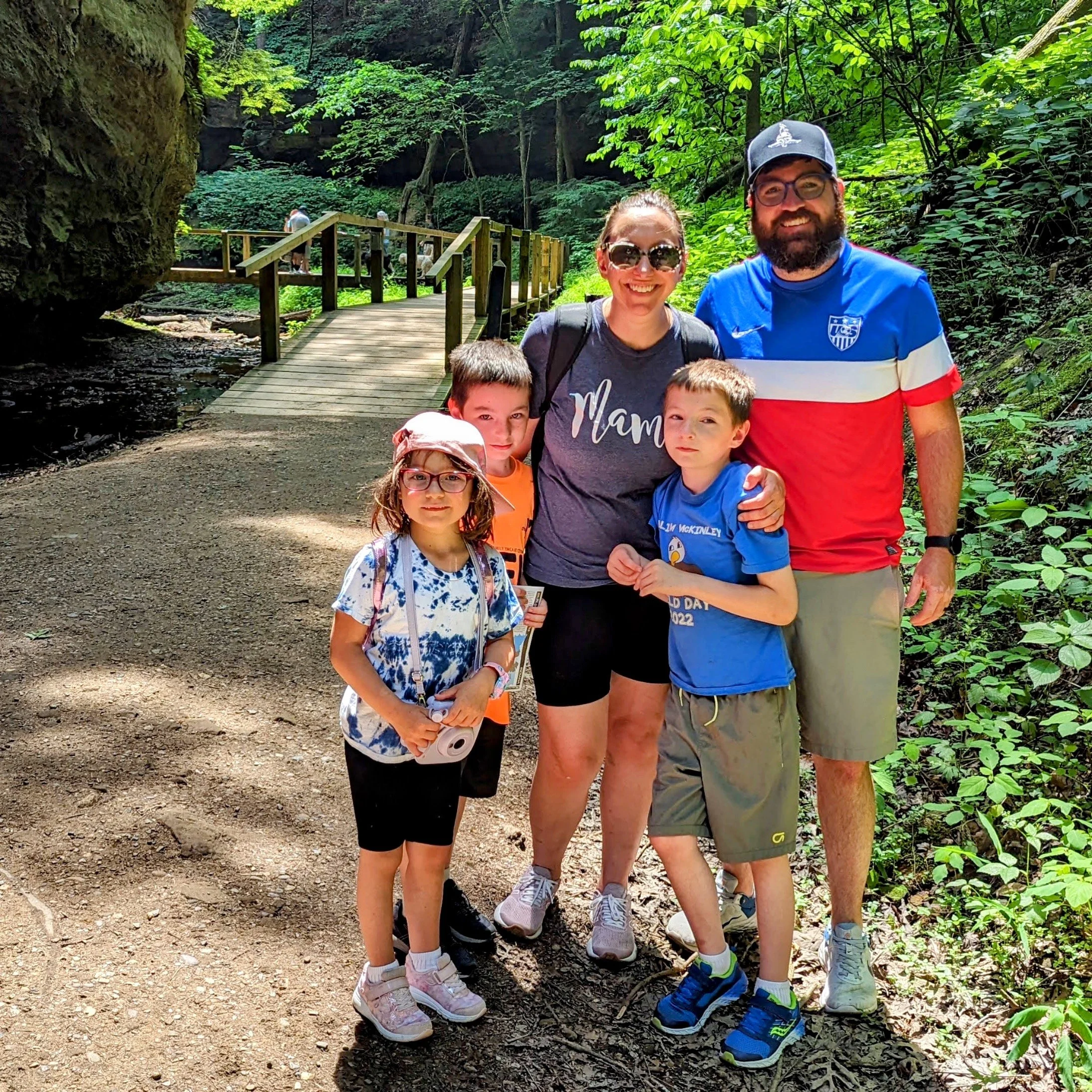 A group of five people, including three children and two adults, standing on a dirt path in a lush green forest with a wooden bridge in the background. They are smiling, wearing casual clothes, and sunglasses, enjoying a day outdoors.