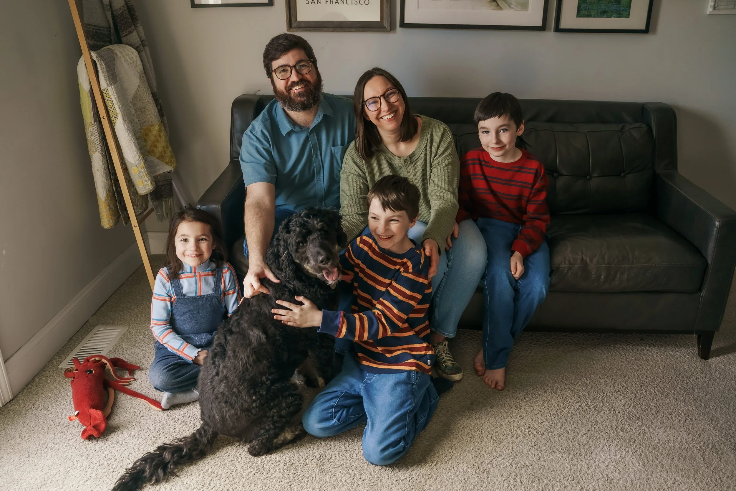 A family of six, including three children, their parents, and a dog, sitting on a black leather couch in a living room. The family is smiling and looking at the camera. There are framed pictures on the wall behind them, and a red plush toy on the carpet. The children are wearing casual clothes, and the dog is a large, curly-haired breed.