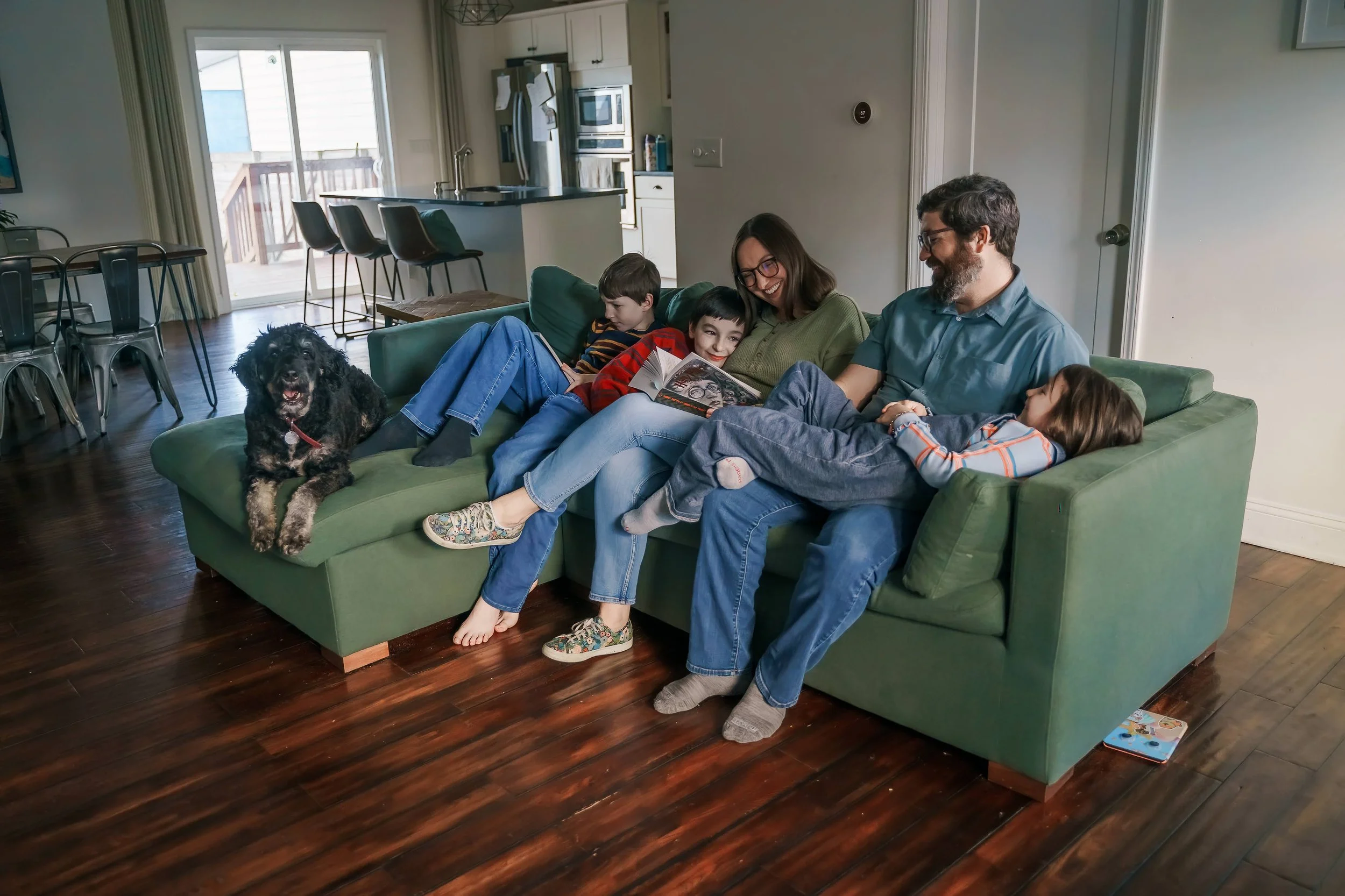 A family of five, including two children, two adults, and a dog, sitting on a green couch in a living room enjoying reading a book together.