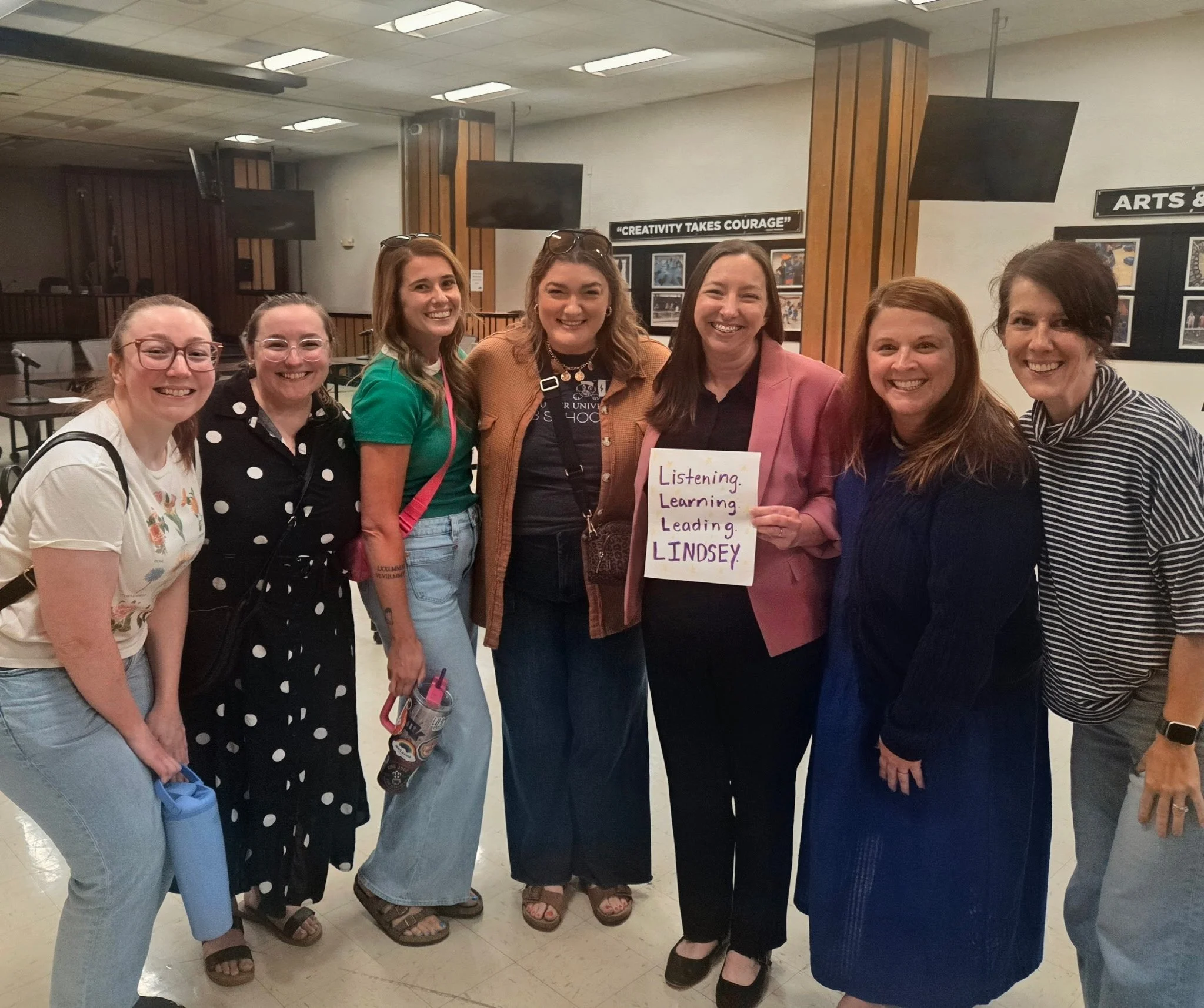 Group of women smiling and holding a sign that says "Listening. Learning. Leading. LINDSEY," in a room with arts and photography displays on the wall.