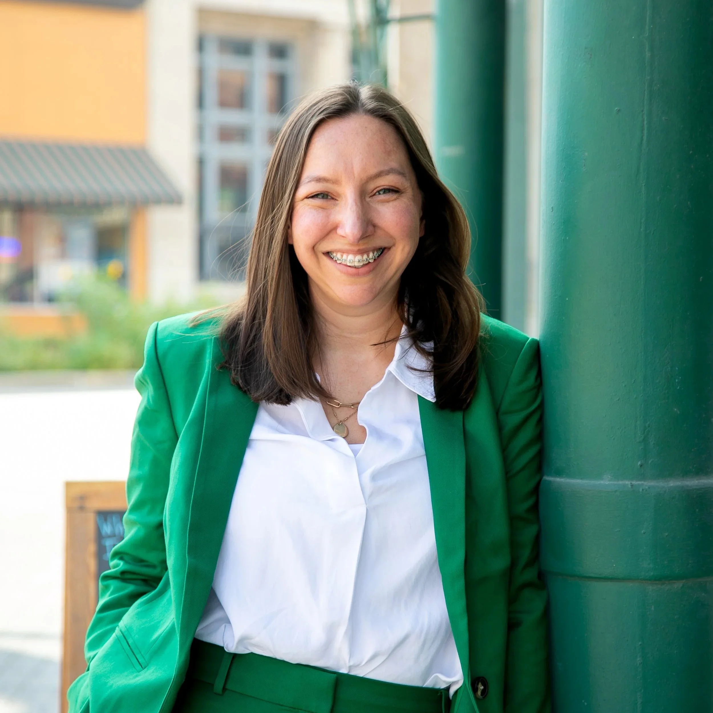 Young woman with long brown hair wearing a white shirt, green blazer, and braces, standing outdoors beside a green pole, smiling at camera.