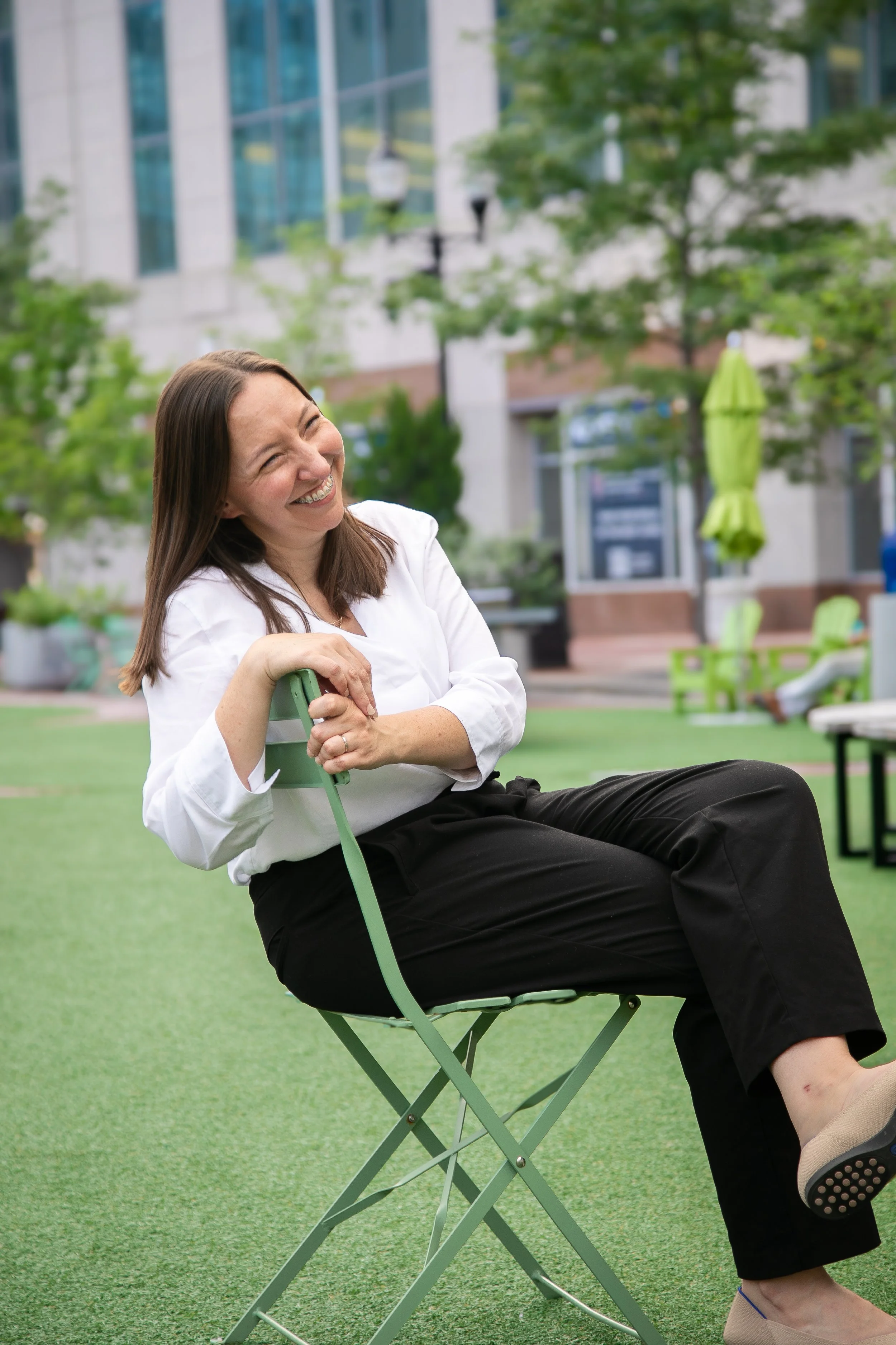 A woman with brown hair and a white shirt sitting on a green folding chair outdoors on an artificial grass surface, smiling and laughing, with a city building, greenery, and outdoor furniture in the background.