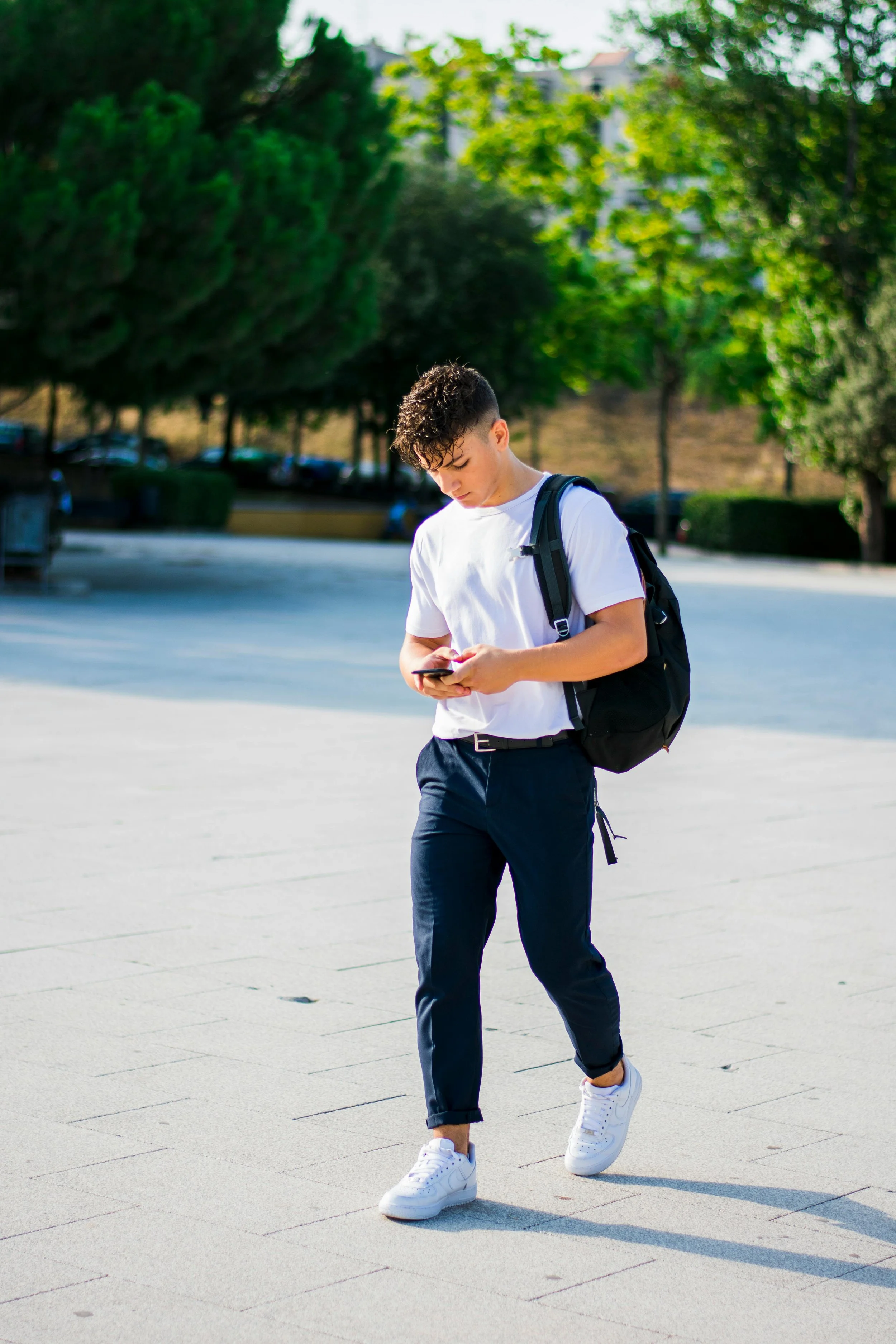 A young man with curly hair wearing a white t-shirt, navy blue pants, white sneakers, and carrying a black backpack, walking outdoors on a sunny day while looking at his phone.
