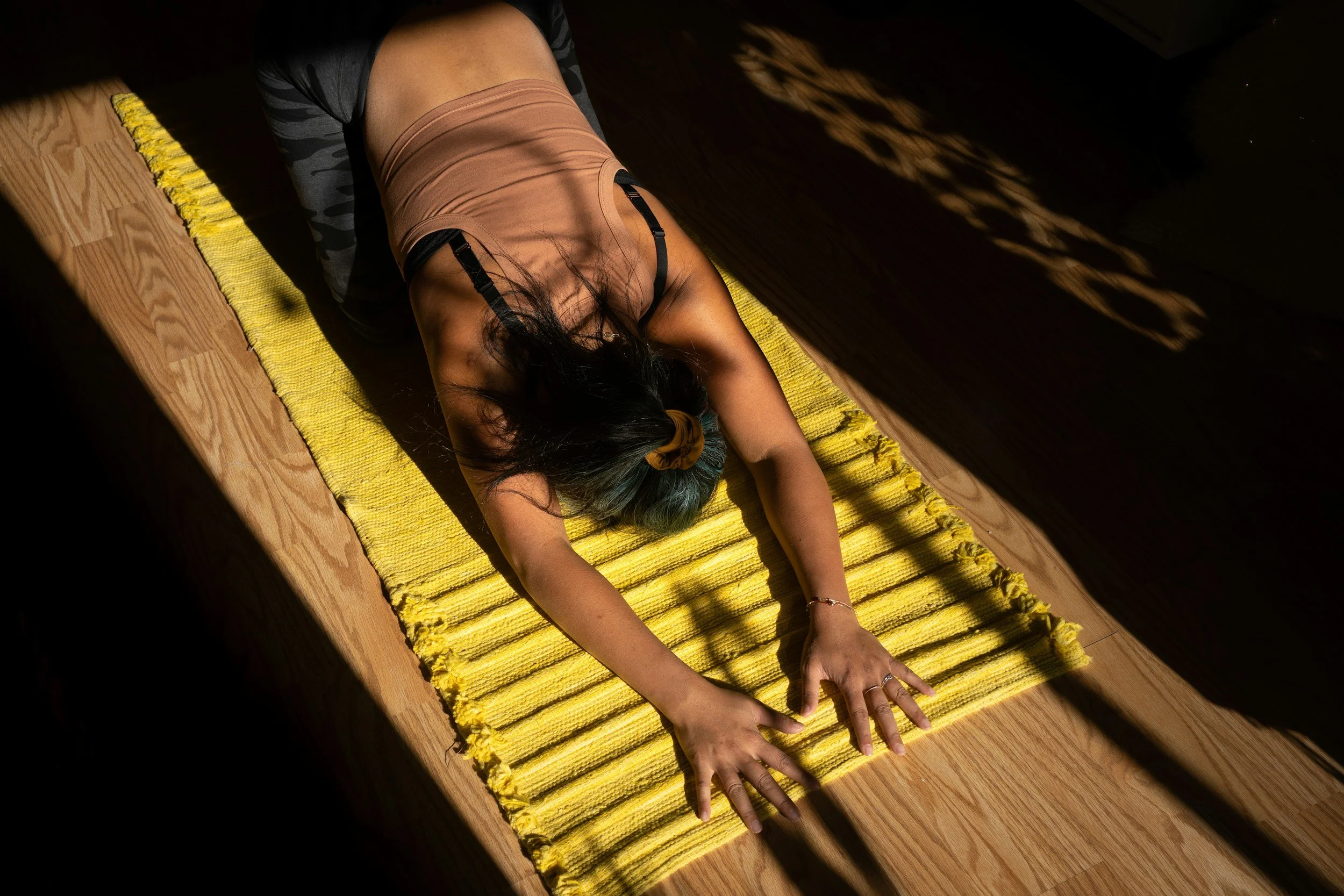 A woman practicing yoga on a yellow mat on a wooden floor, lying face down with her arms extended forward, hair covering her face, in a dimly lit room with shadows.