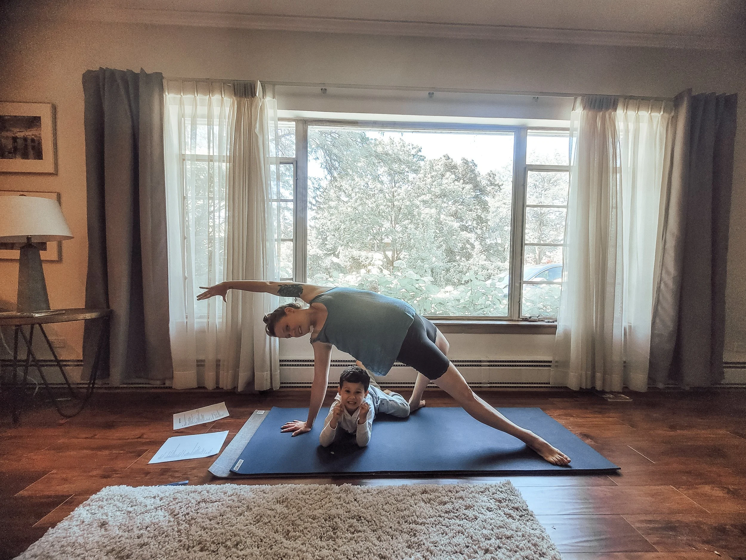 A woman doing yoga on a blue mat in a room with large window and white curtains, with a young boy lying underneath her on the mat, smiling and pointing at the camera.