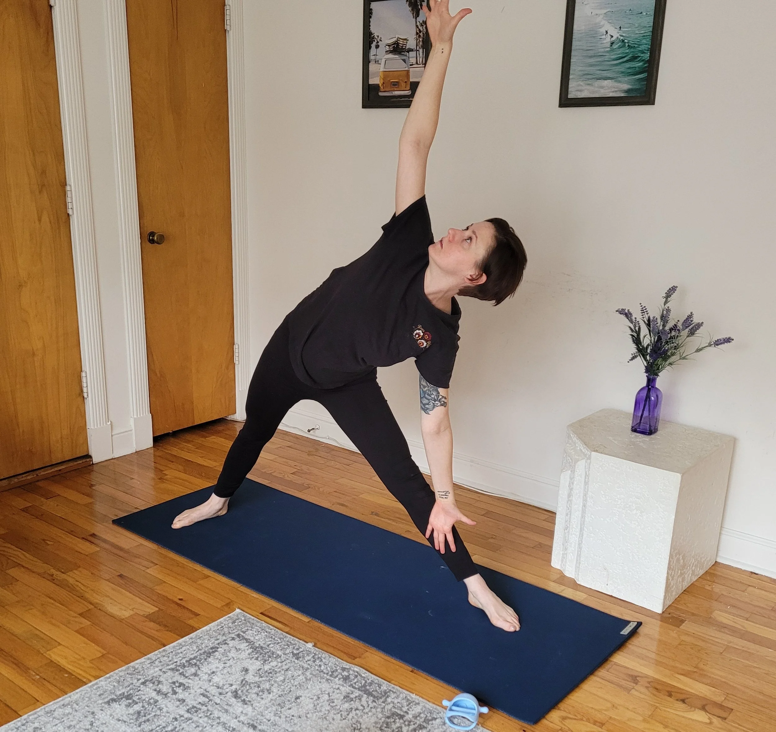 A woman practicing yoga at home, standing on a black exercise mat, wearing a black shirt and black pants, with a tattoo on her left arm, in a room with wooden floors, white walls, and framed pictures of beaches and waves. A purple vase with flowers is on a small white table nearby, and a blue pair of slippers is on the floor next to the mat.