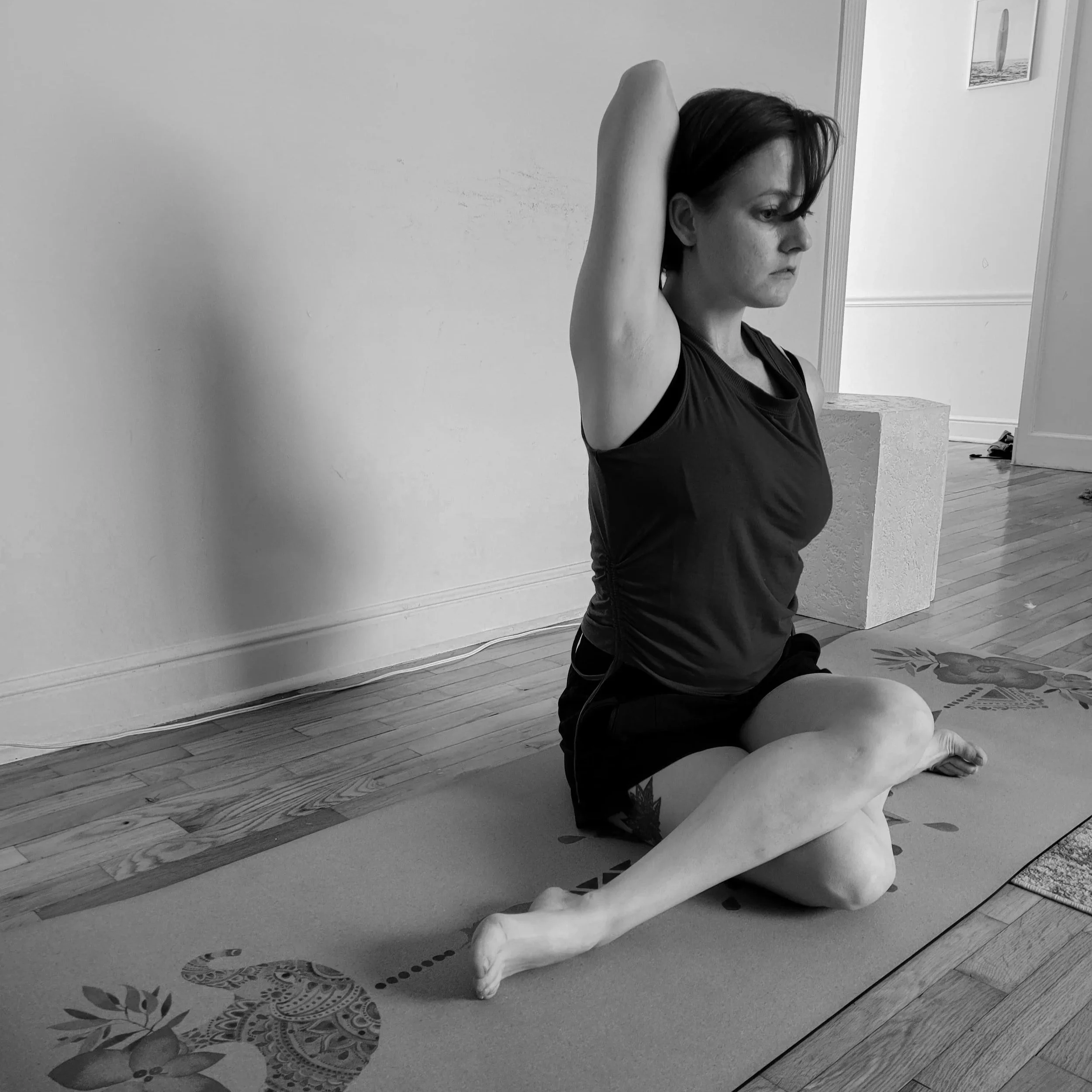 A black and white photograph of a woman sitting on her knees on a yoga mat, practicing a yoga pose with her arms raised behind her head, in a room with a plain wall, framed pictures, and hardwood floors.