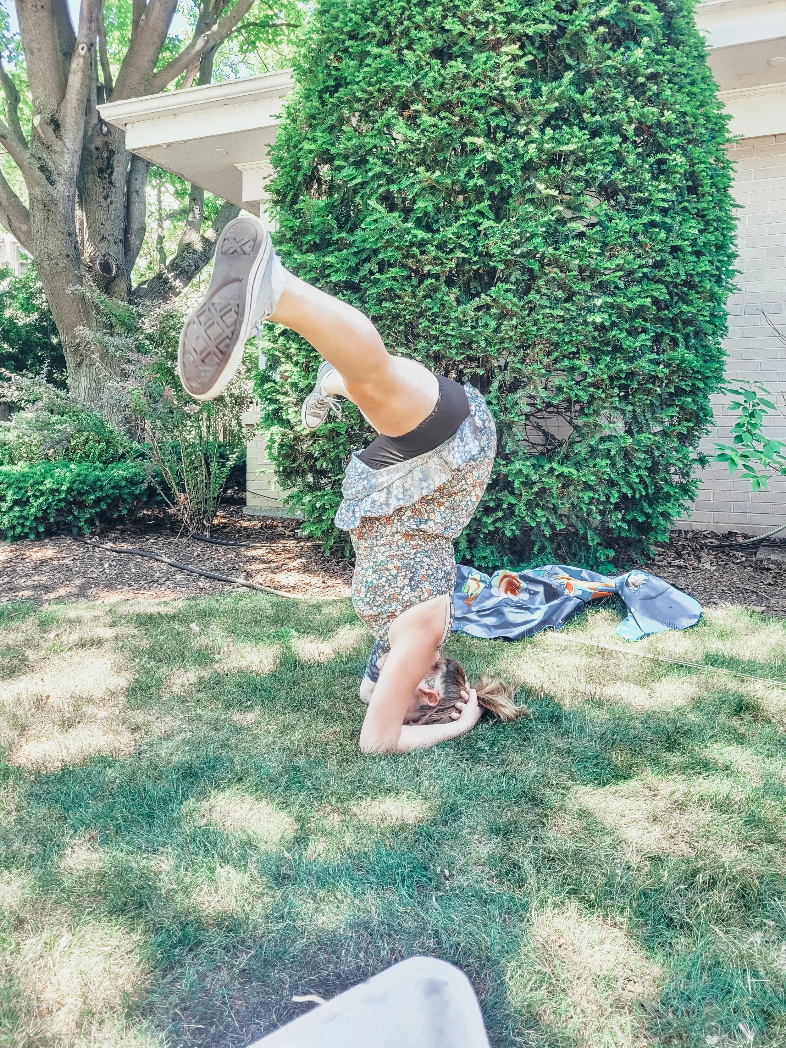 A person performing a handstand outdoors on green grass, with trees and shrubbery in the background, seen from the perspective of someone sitting on the ground.
