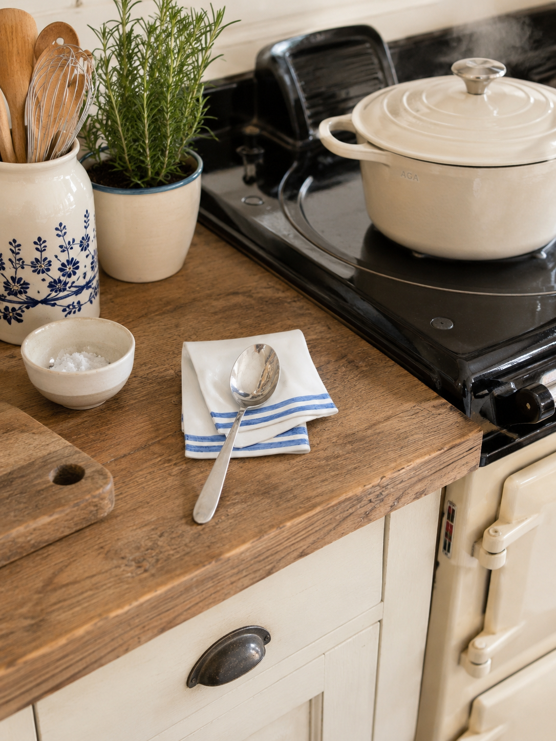A rustic kitchen counter with a wooden cutting board, salt in a small bowl, a white cloth napkin with blue stripes, a silver spoon, potted herbs, and a stove with a steaming white pot.