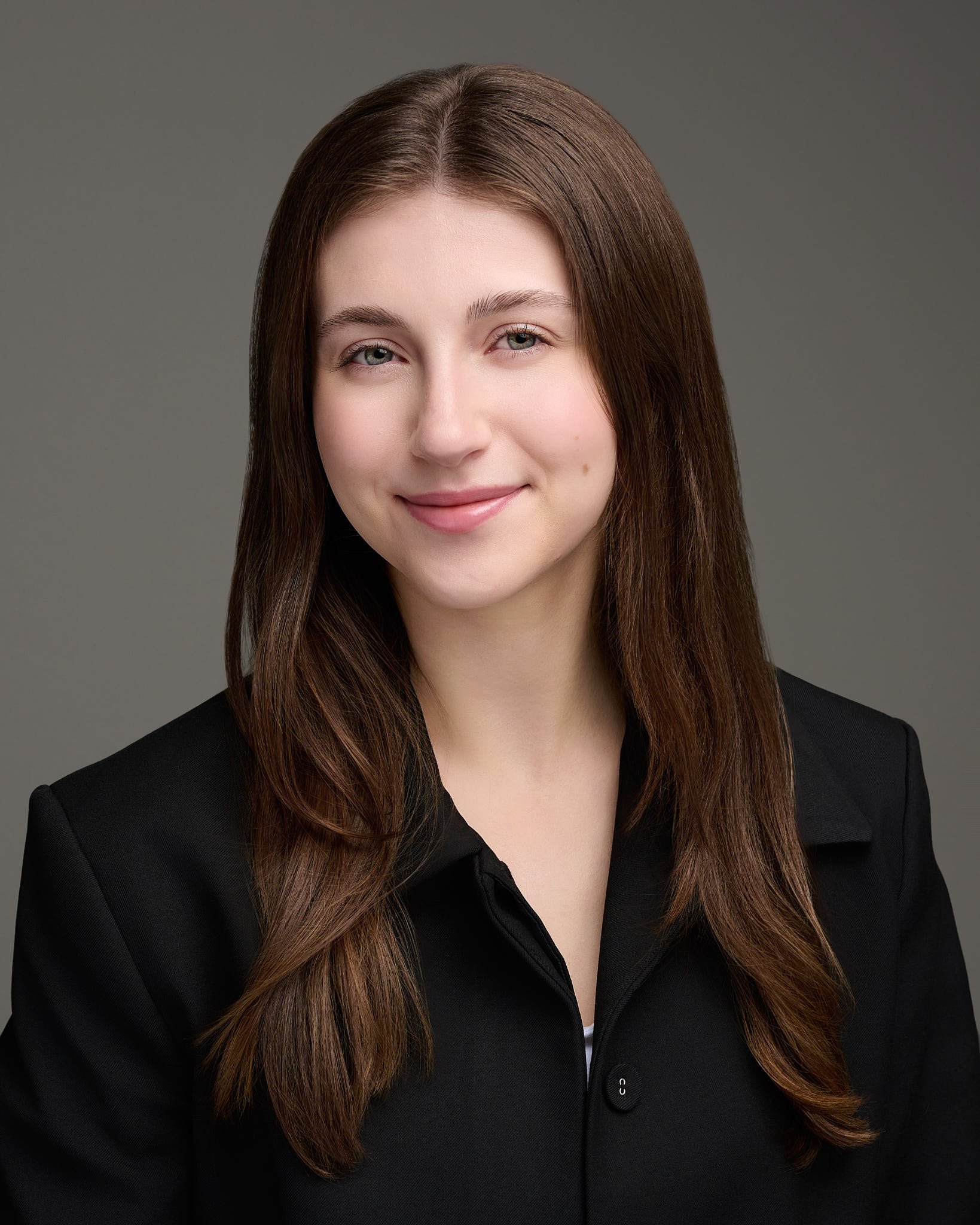 A young woman with long brown hair and light skin, wearing a black blazer, smiling confidently at the camera against a plain gray backdrop.