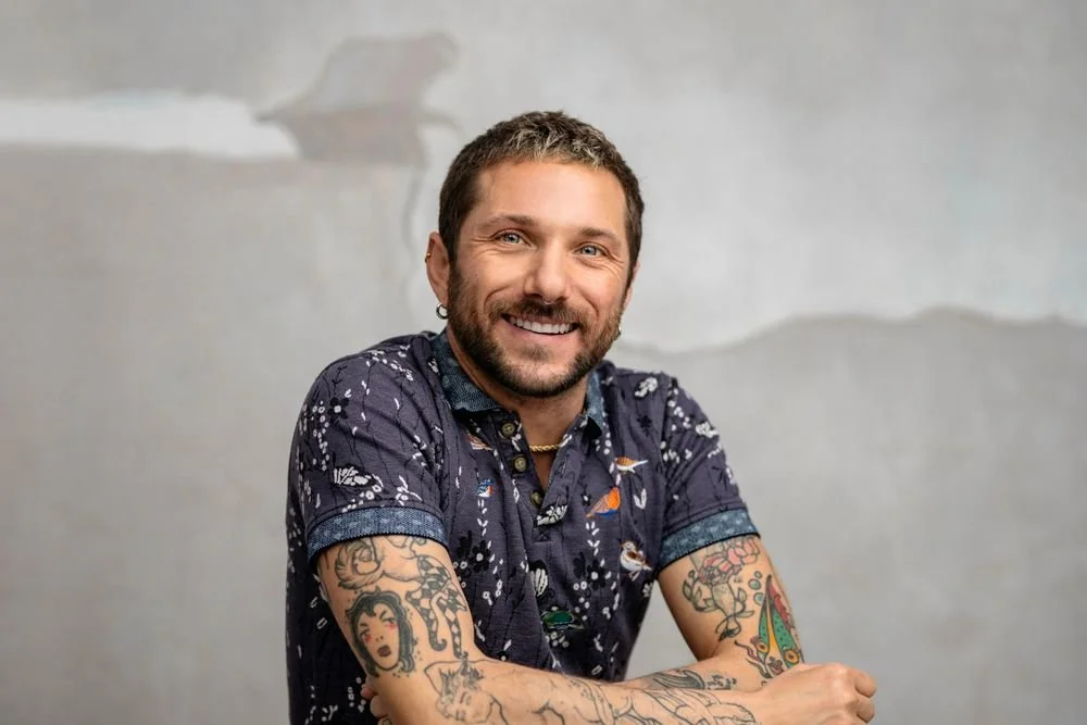 Smiling man with tattoos on his arms, wearing a dark patterned shirt, seated against a plain, textured wall.