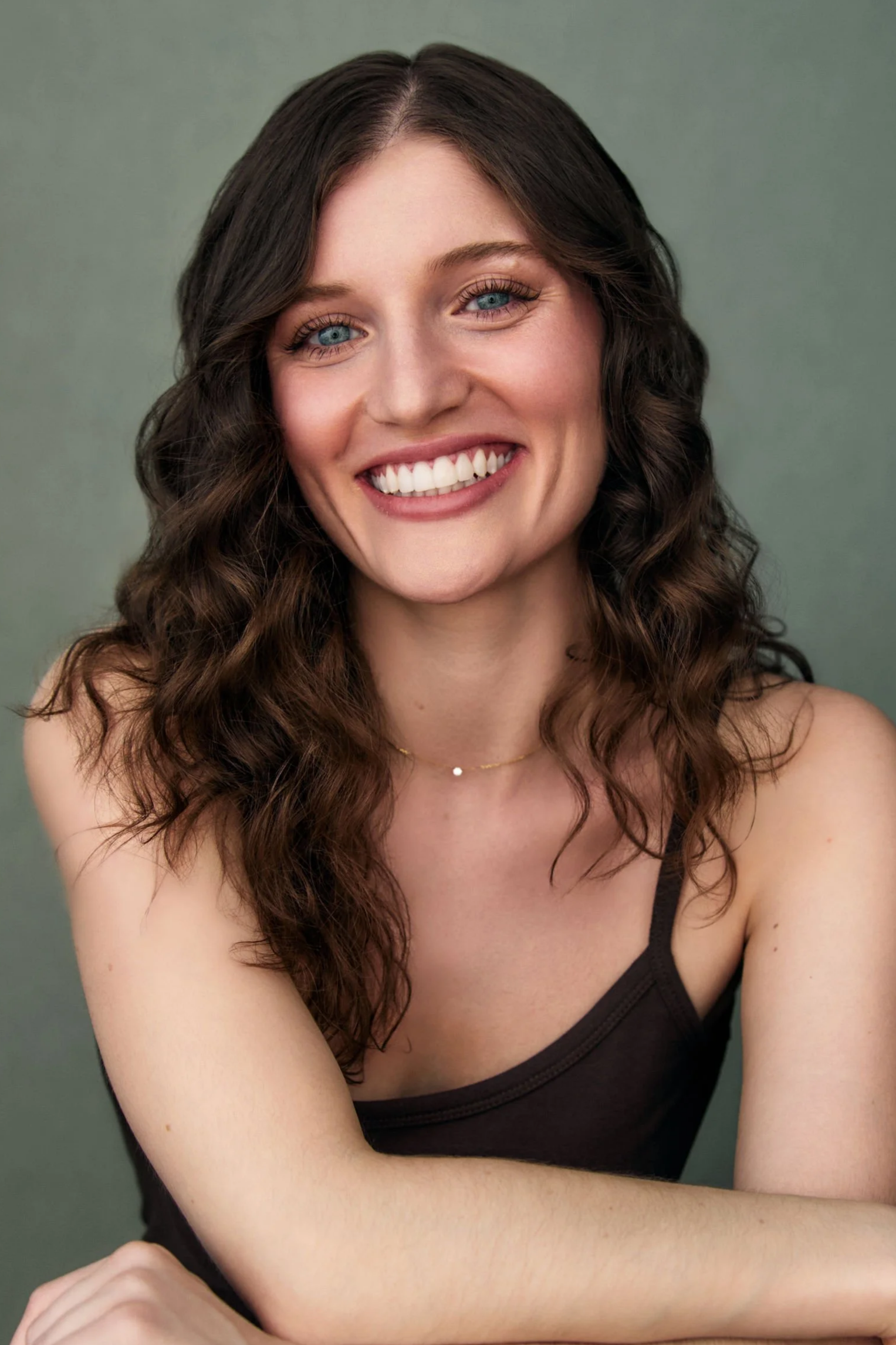 Close-up of a smiling woman with blue eyes, long curly brown hair, wearing a black tank top and a delicate necklace, posed against a muted green background.