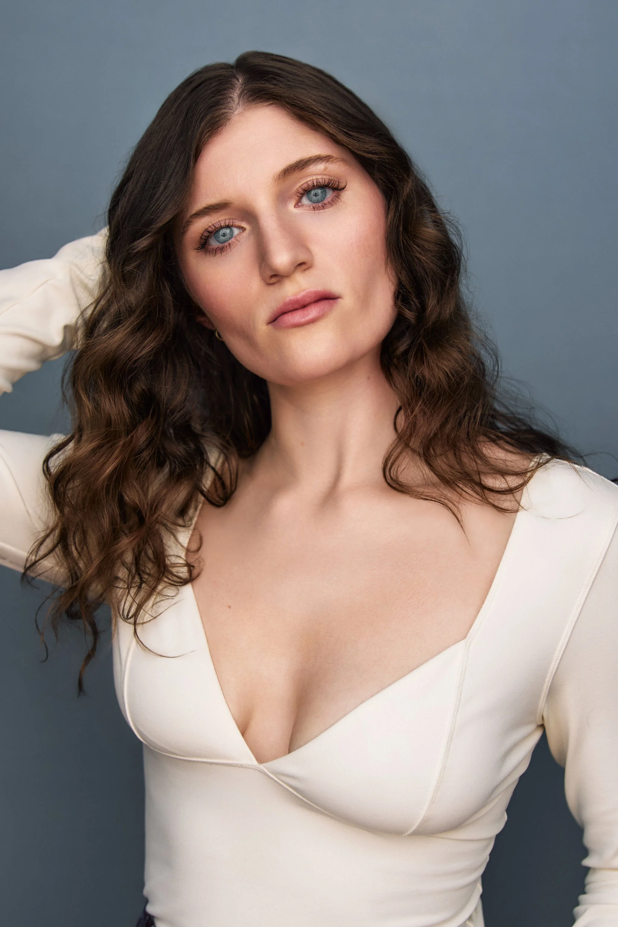 A woman with long wavy brown hair, blue eyes, wearing a white V-neck top, posing with her hand behind her head against a gray background.