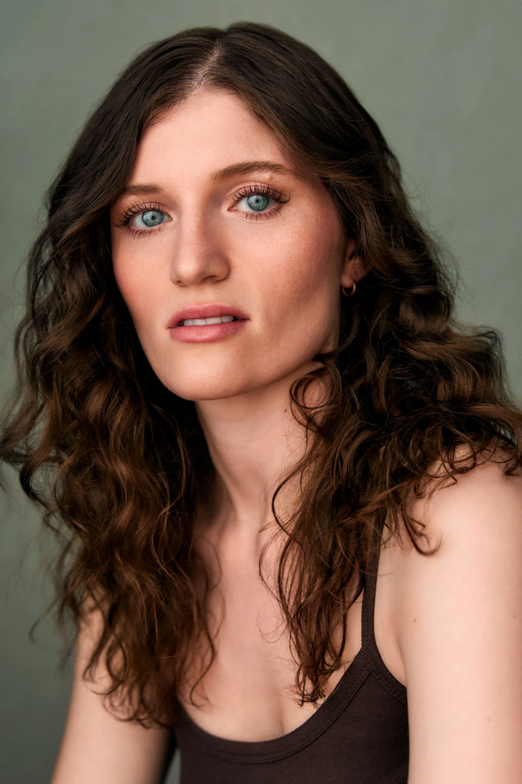 Close-up of a woman with curly brown hair and blue eyes wearing a black tank top, against a green background.