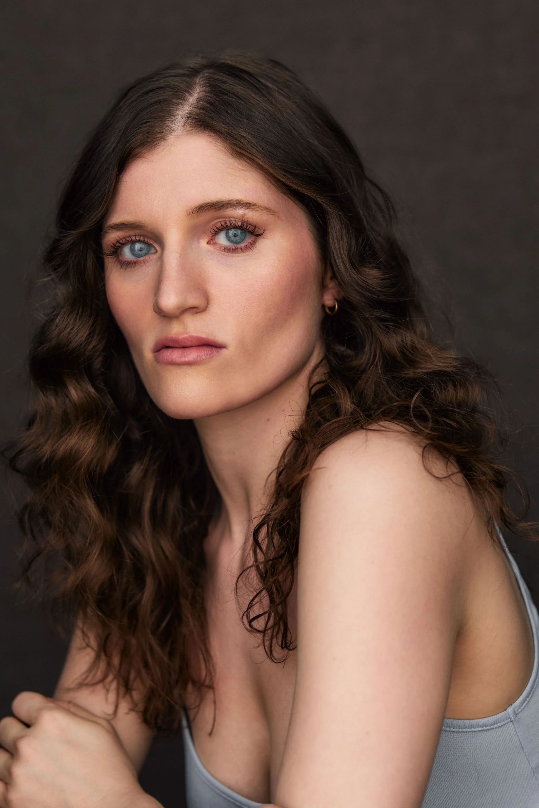 Close-up of a young woman with curly brown hair, blue eyes, and light skin, wearing a gray tank top, looking directly at the camera against a dark background.
