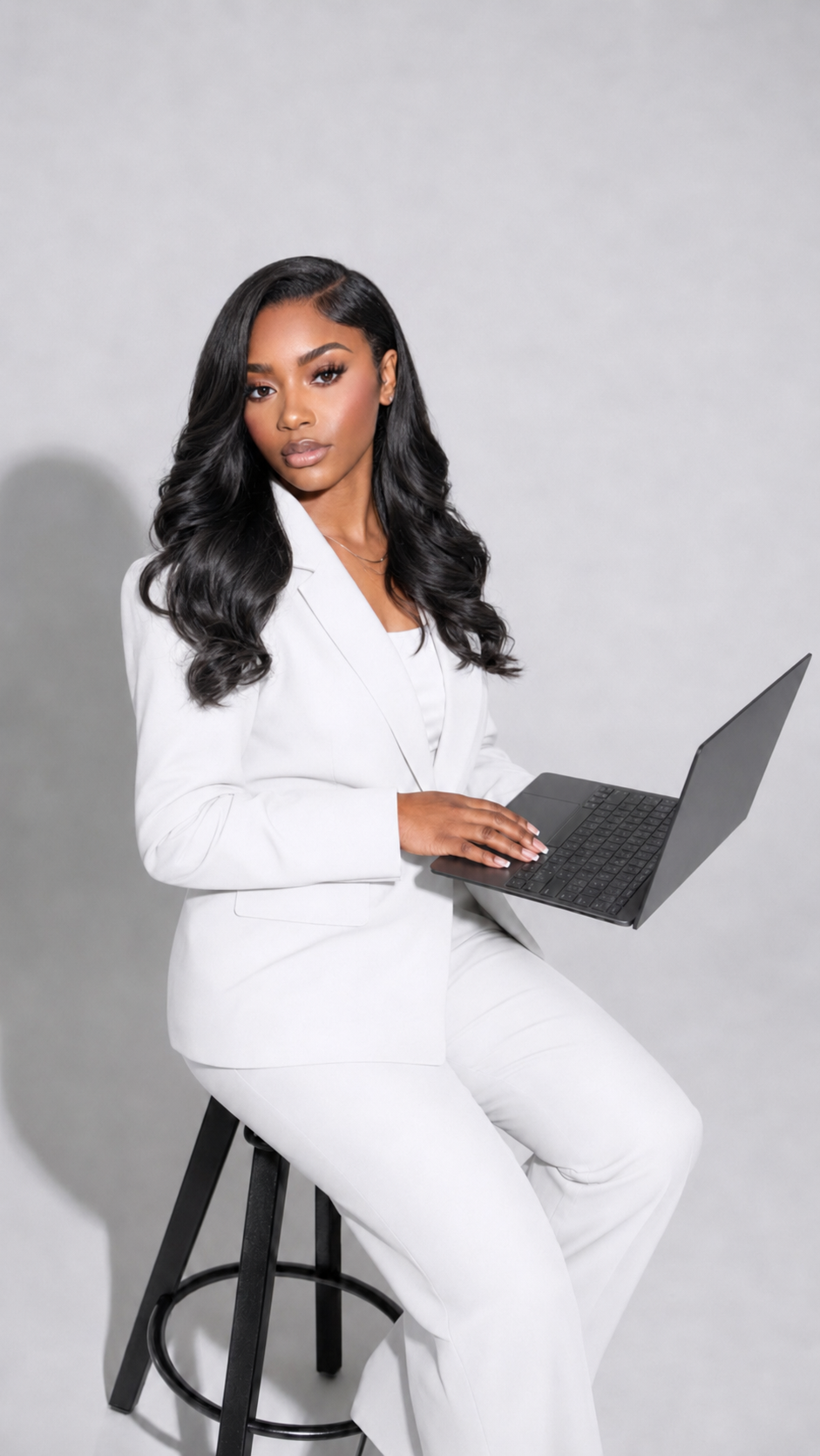 A woman in a white business suit sitting on a black stool, holding a laptop.