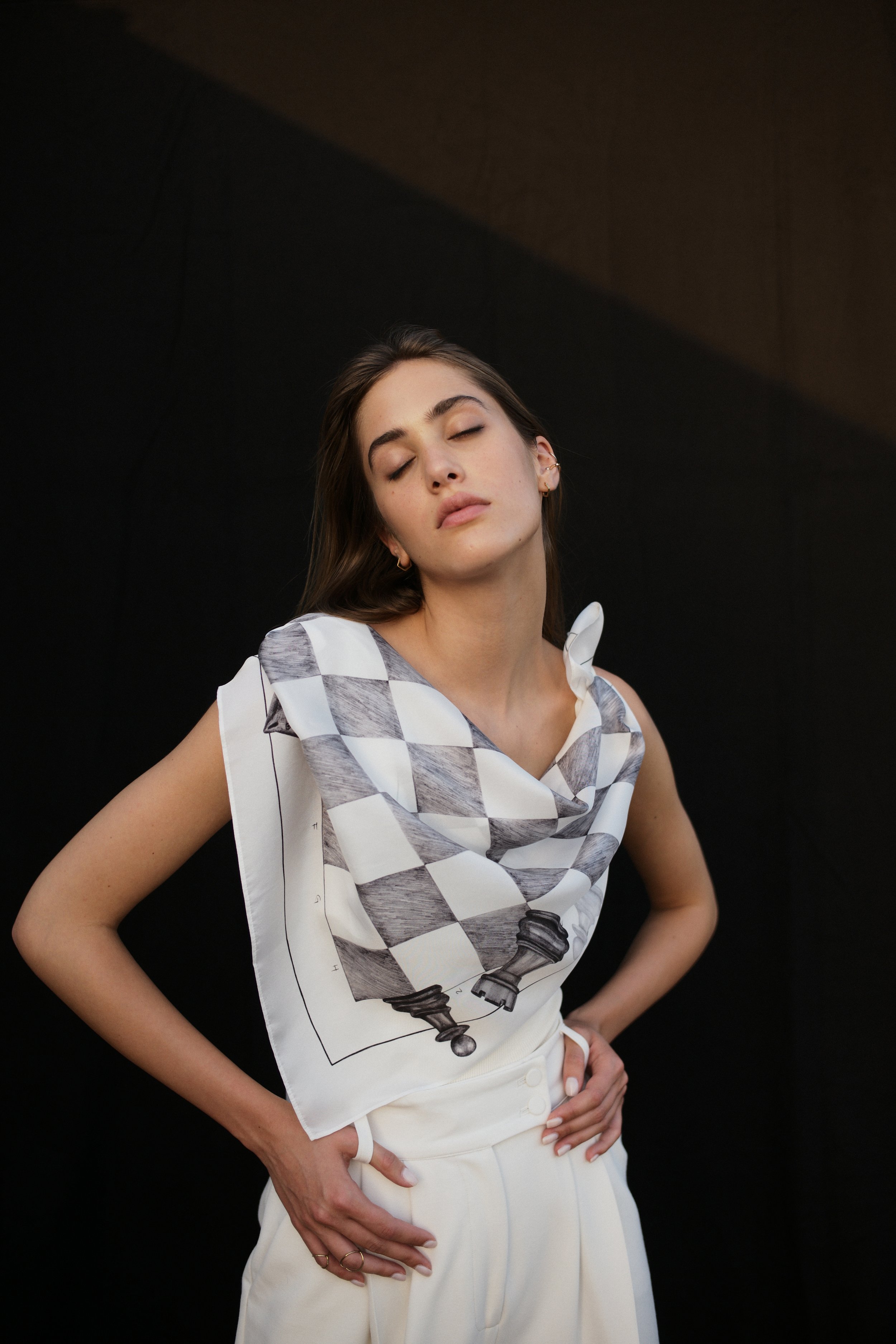 A young woman with long brown hair and closed eyes, wearing a white sleeveless top with a chessboard pattern, posing against a dark background.