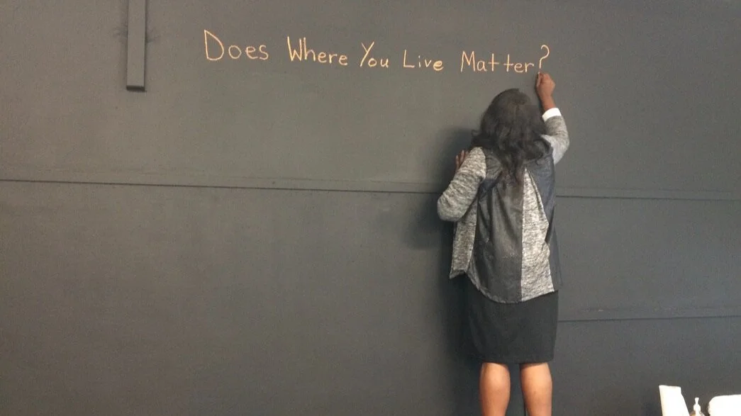 A woman writing the question "Does where you live matter?" on a dark gray wall with orange chalk on her class's learning wall.
