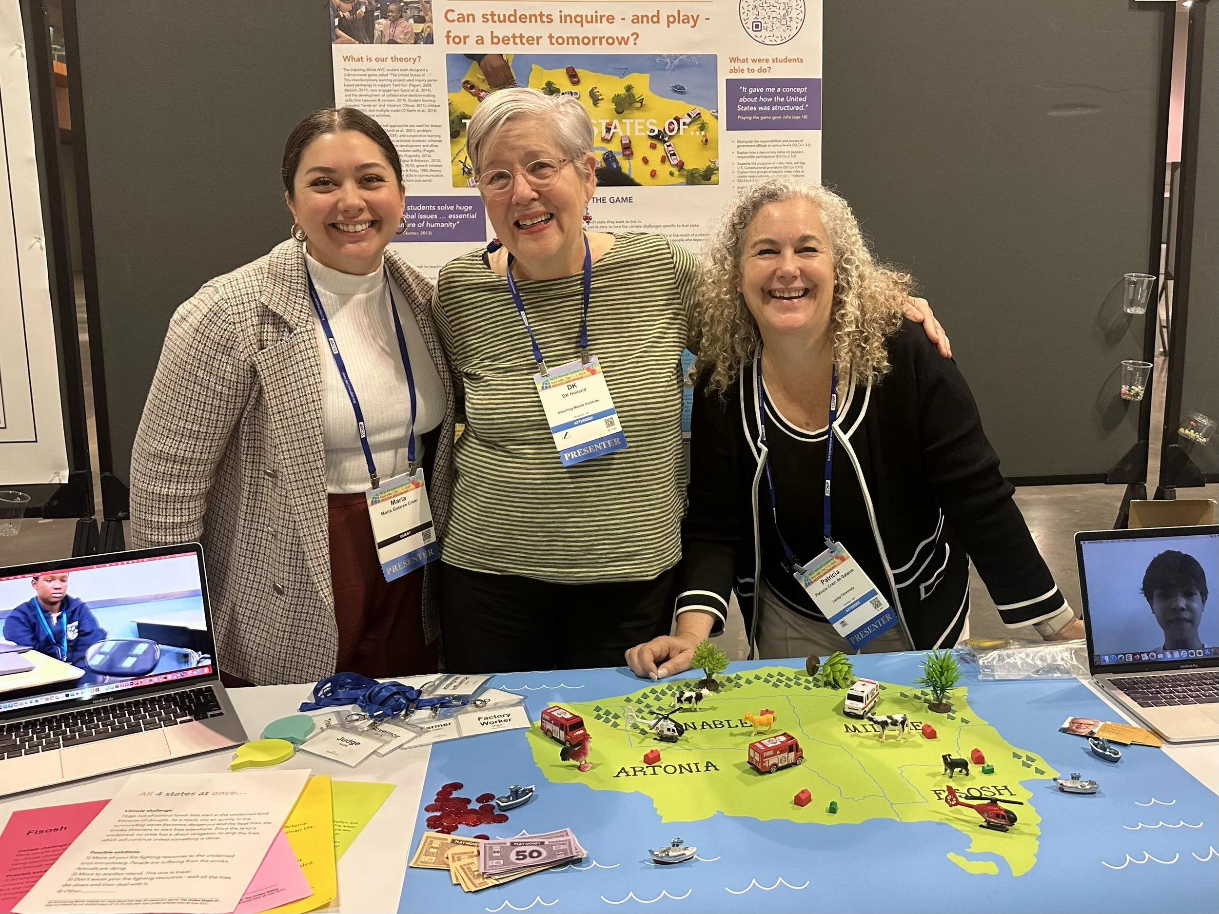 DK Holland and Patricia Crain de Galarce smiling and standing behind a table at a NCSS conference, displaying a colorful map of the "United States of..." with small toy models of farms, buildings.