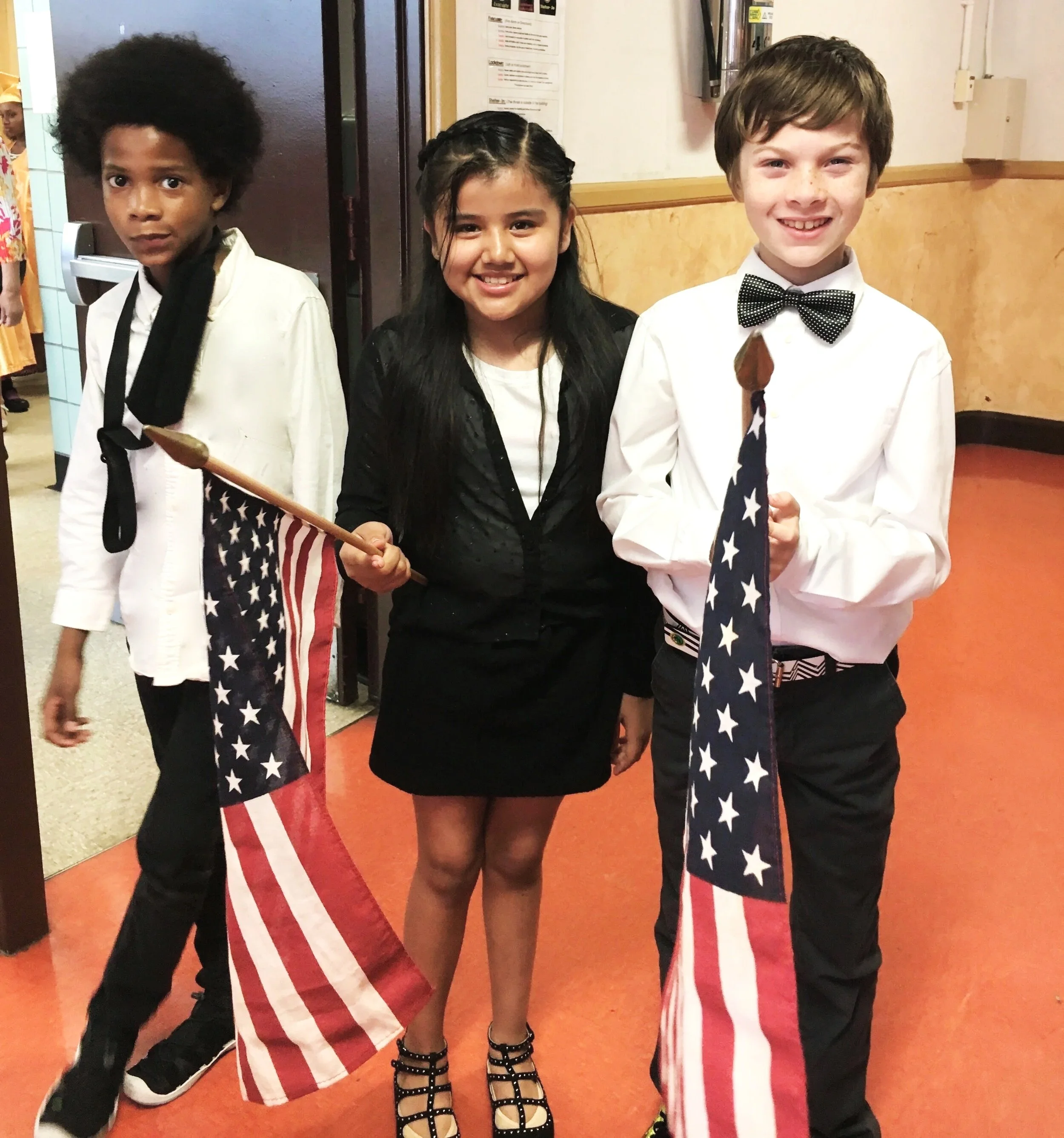 Three children standing in a hallway, holding American flags. The boy on the right is wearing a white shirt with a black bow tie and black pants. The girl in the middle has long dark hair and is dressed in black. The boy on the left has curly hair, wearing a white shirt with a black tie. They are smiling and appear to be celebrating or participating in a patriotic event.