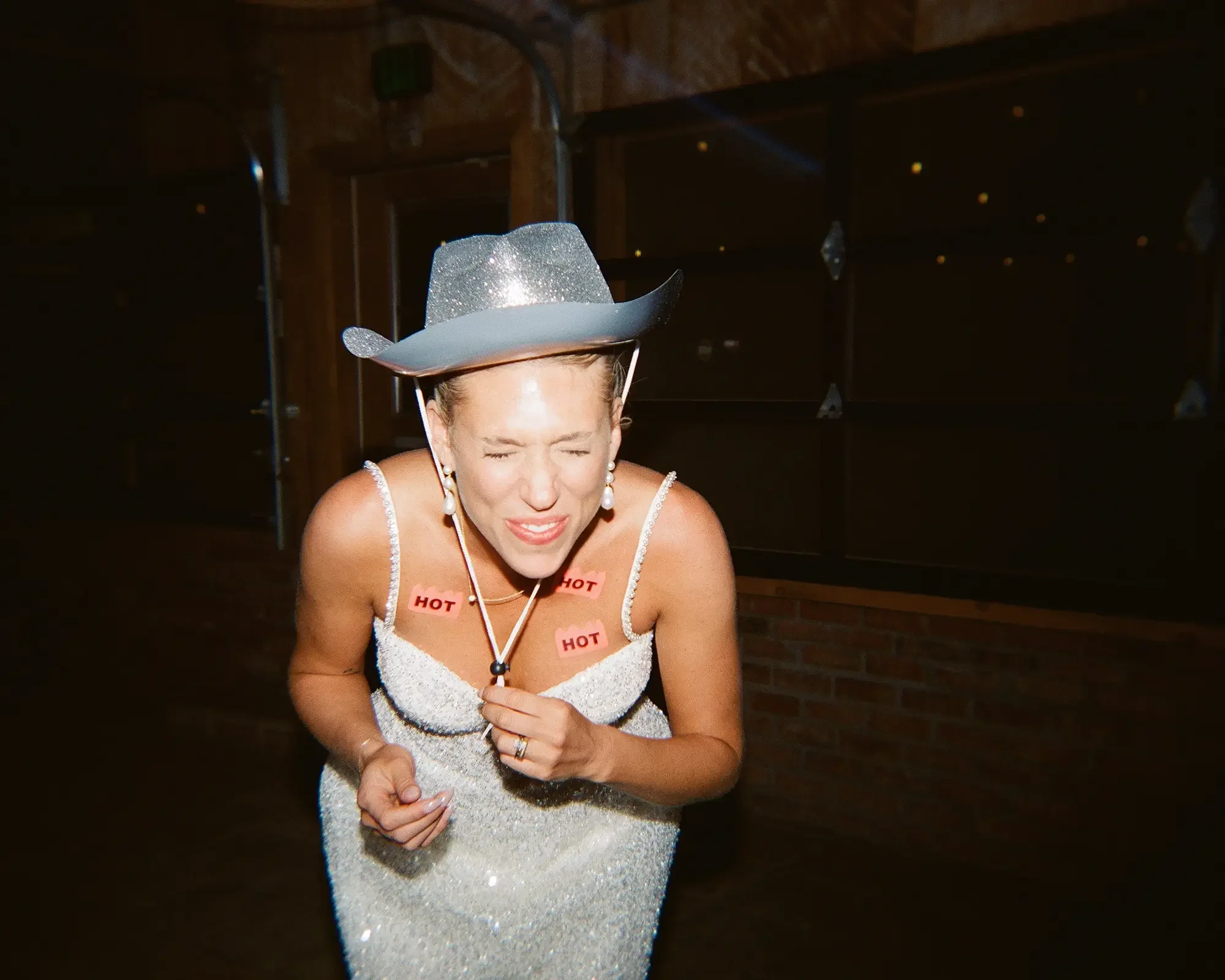 A woman wearing a sparkly dress and a glittery cowboy hat, with 'HOT' stickers on her chest, appears to be tired or in discomfort in an indoor setting with dark walls and musical equipment in the background.
