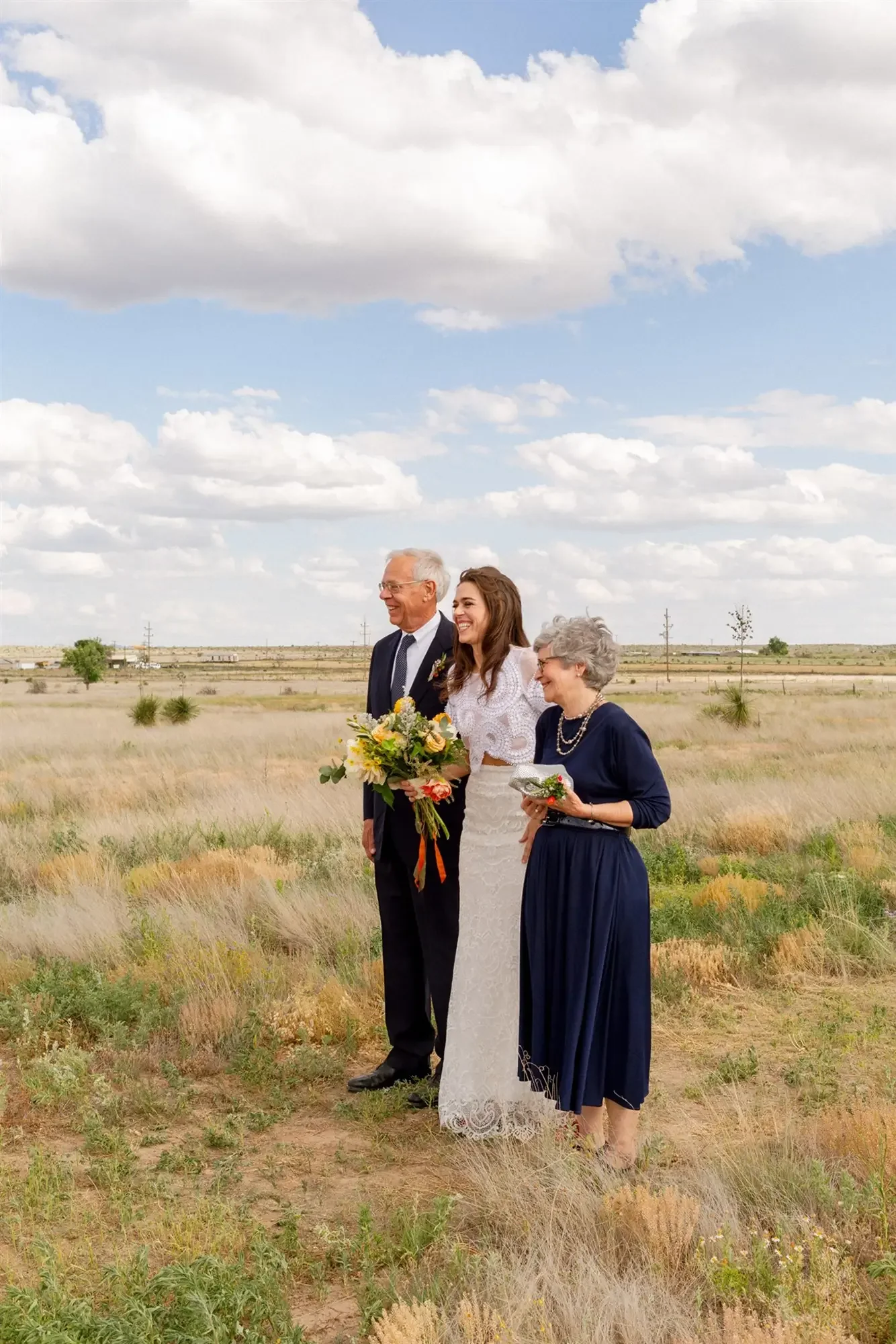 The bride and her parents are standing outdoors in a grassy field under a partly cloudy sky before the ceremony, with the bride in the middle wearing a white lace dress, holding a bouquet of flowers. 
