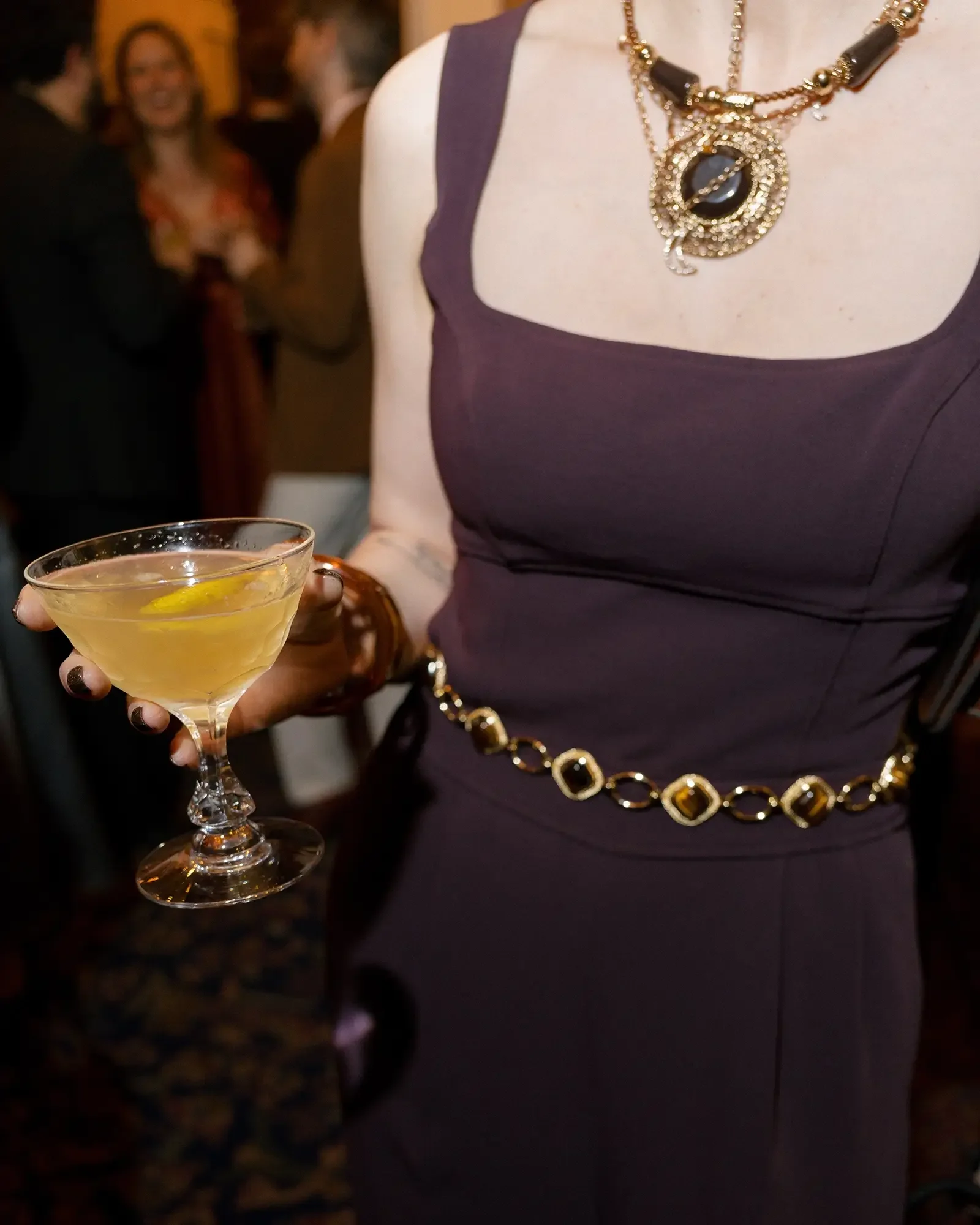 A woman in a purple dress holding a martini glass with a yellow drink and lemon slice, wearing a gold necklace and belt with large gemstones, at a social event with blurred people in the background.