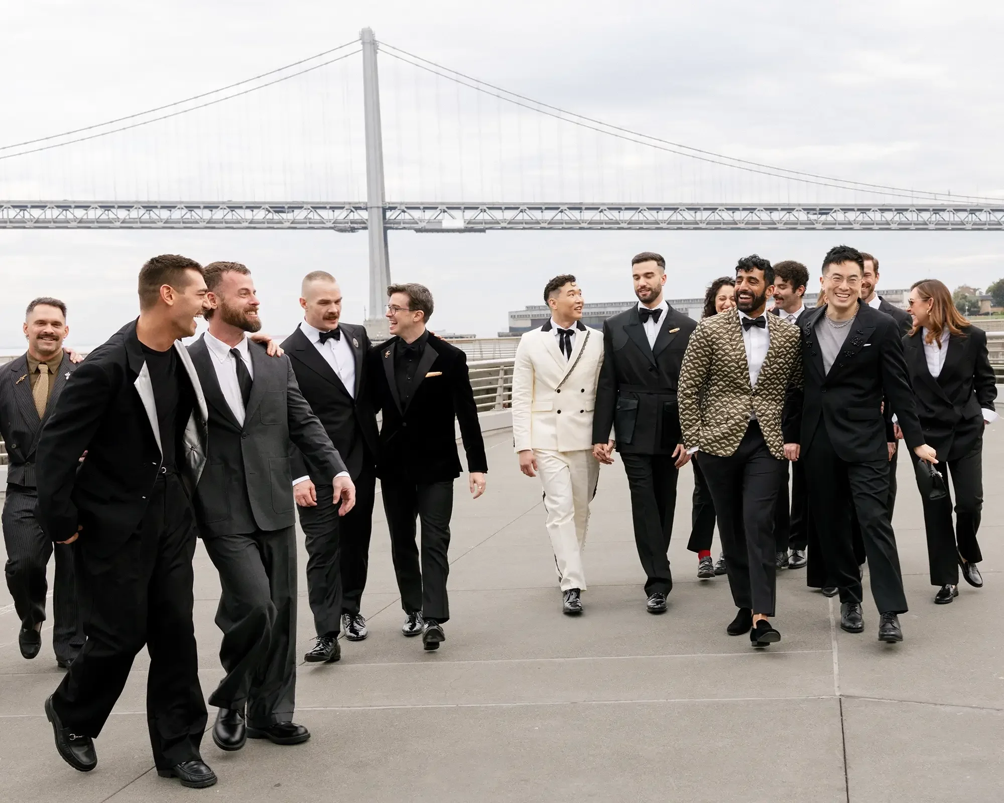 Wedding party dressed in tuxedos and suits walking outdoors near the bay bridge, smiling and engaging in conversation.