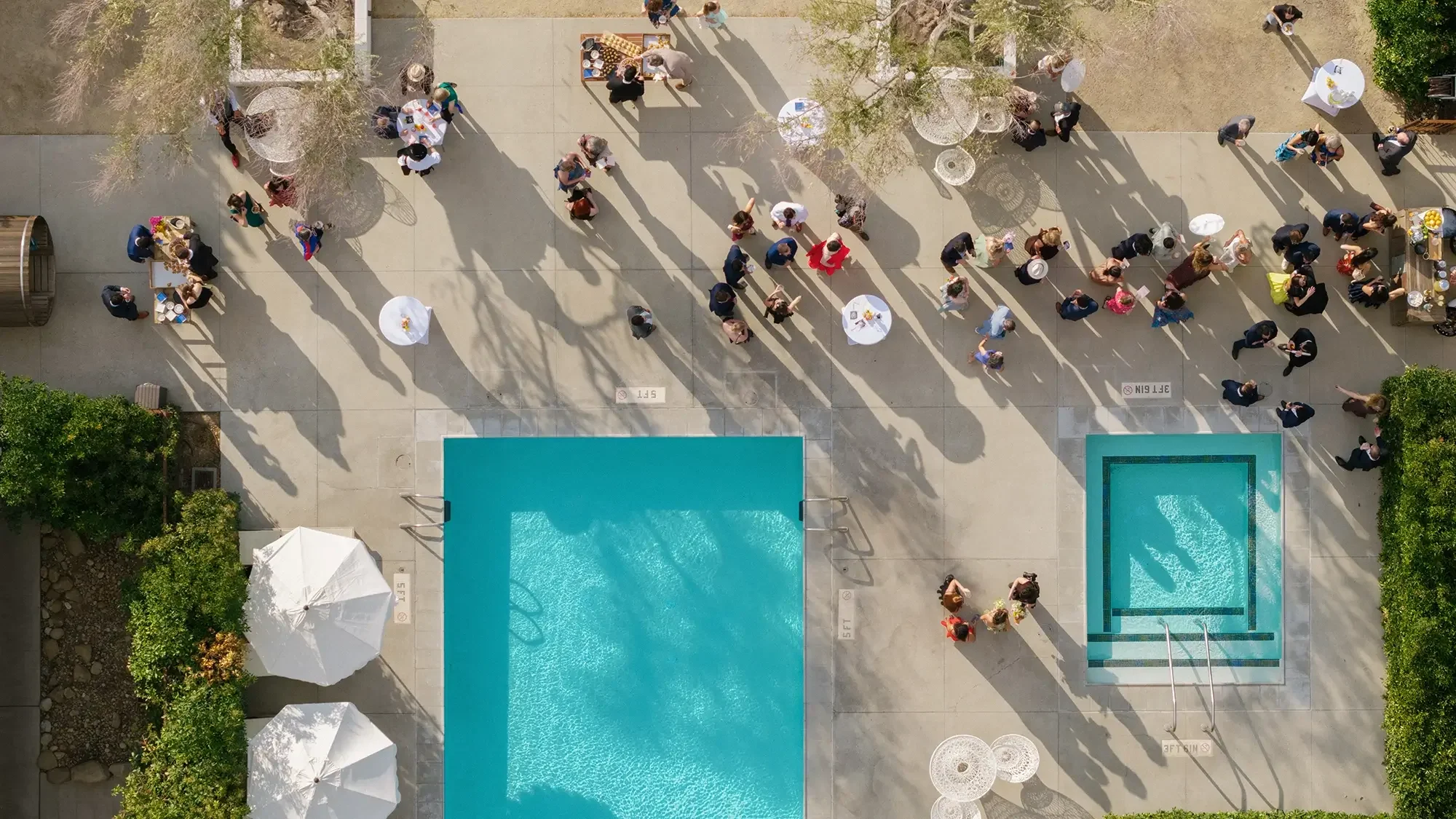 People gathered around a swimming pool and a hot tub on a sunny day, with shadows cast by trees and umbrellas during a wedding cocktail hour