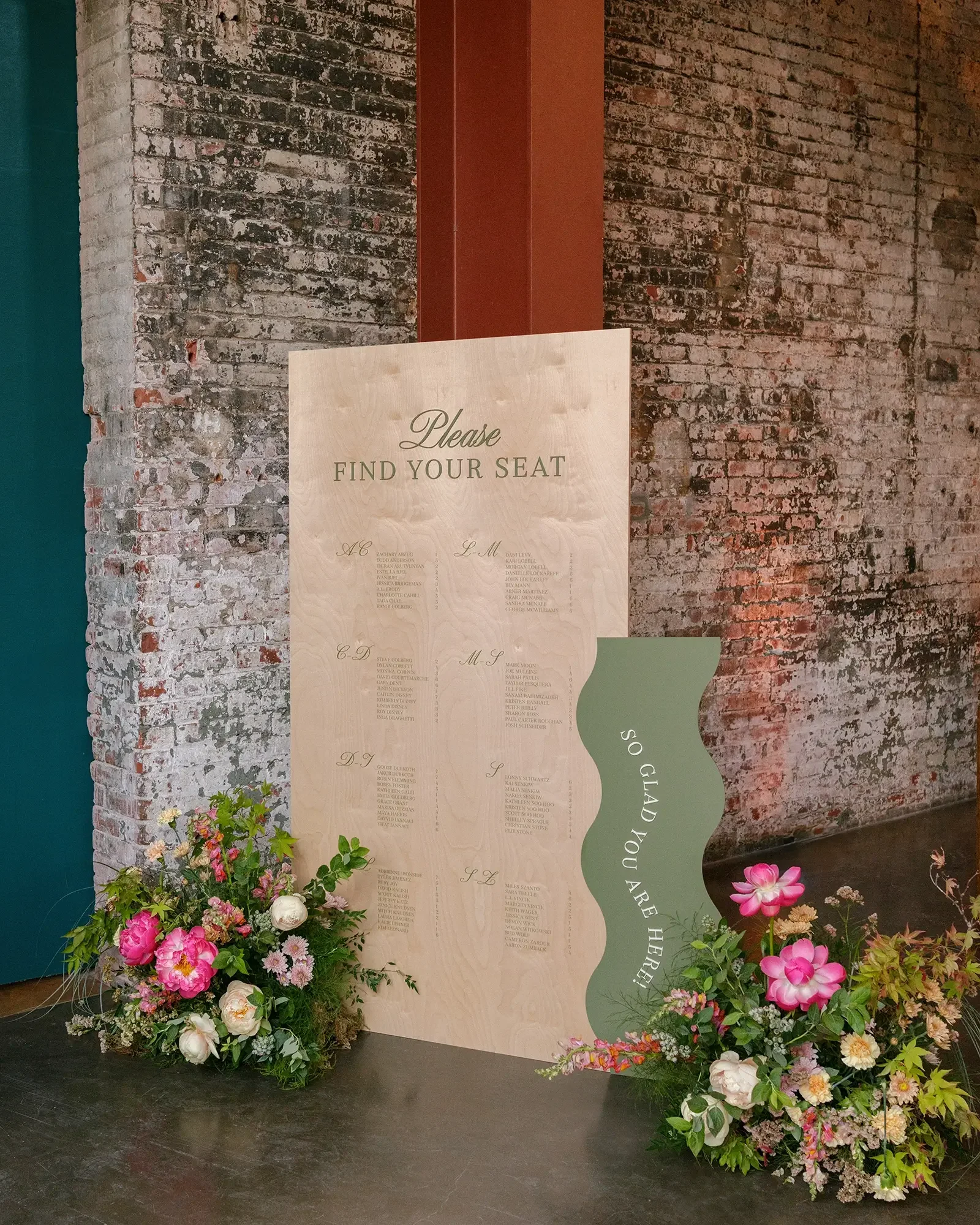 A wooden wedding seating chart board with floral arrangements at the base, positioned against a brick wall. The board has a green wavy side with text 'So glad you are here' and lists of guests organized by table.