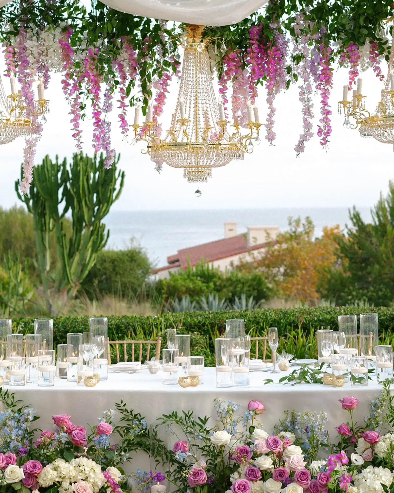 Outdoor wedding reception table with floral arrangements, glassware, and chandeliers, overlooking a landscape with trees, bushes, and a distant body of water.