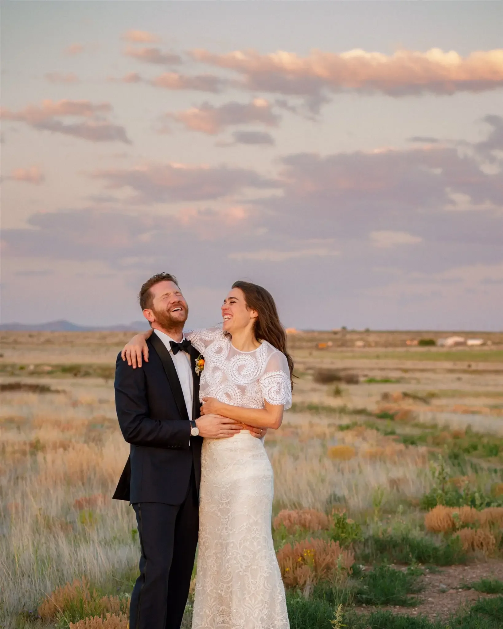 A happy couple in wedding attire, a man in a tuxedo and a woman in a white lace wedding dress, laughing and embracing outdoors in a field at sunset