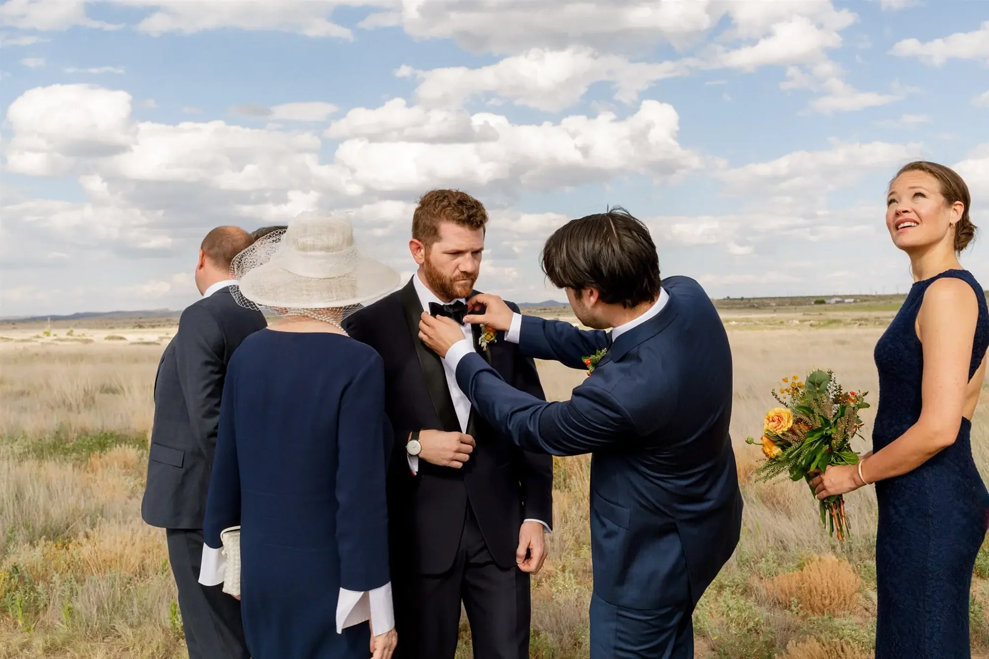 A wedding ceremony taking place in Marfa, TX outdoors in a field with five people. A man is adjusting a bow tie on the groom, while a woman in a navy blue dress is holding a bouquet, looking upward and smiling.