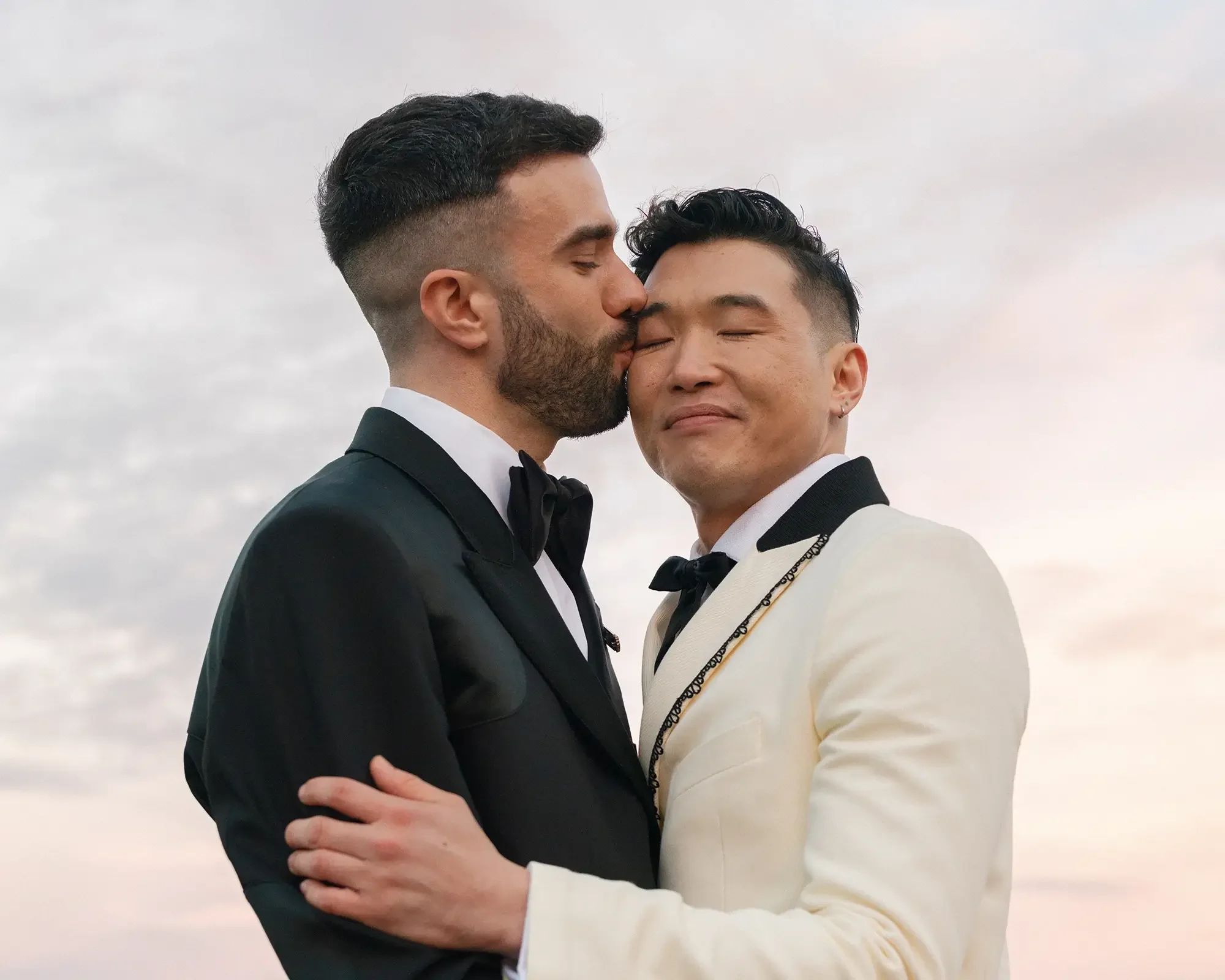Two men in tuxedos embracing and kissing each other at sunset, one in a black tuxedo and the other in a white tuxedo.