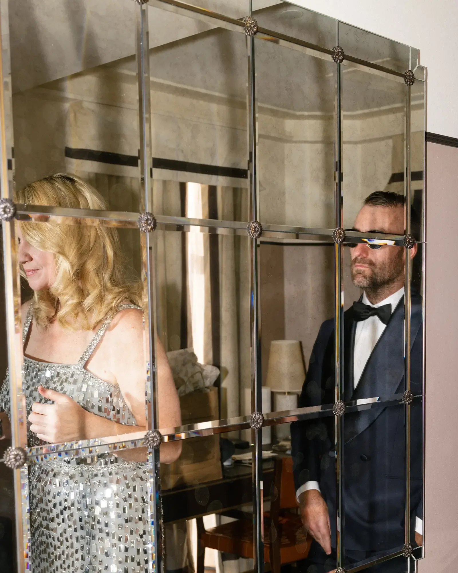 A bride in a silver dress and a groom in a black tuxedo and sunglasses stand behind a mirrored partition in a room.
