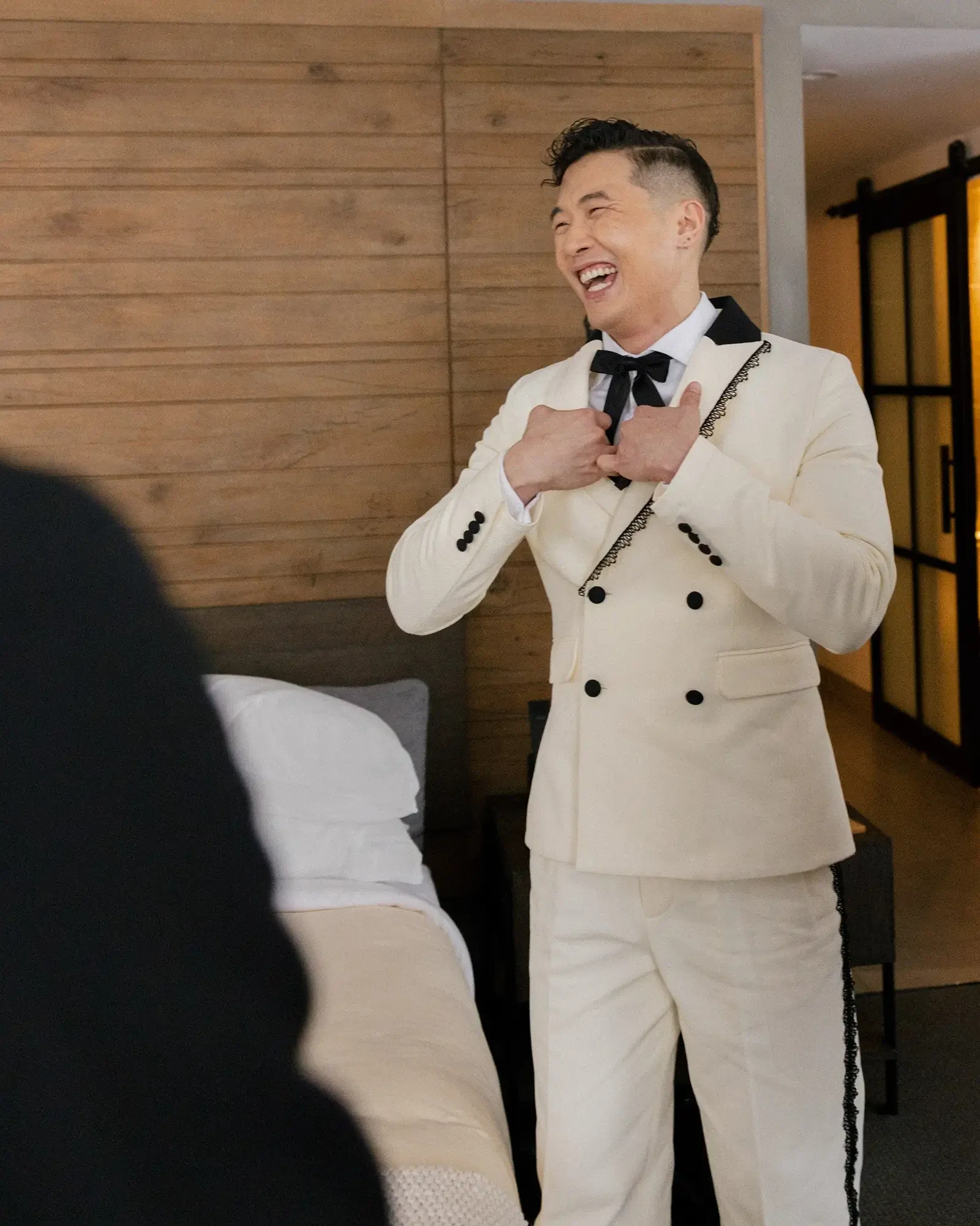 Joel Kim Booster in a white tuxedo with black accents laughing and adjusting his bow tie in a bedroom.