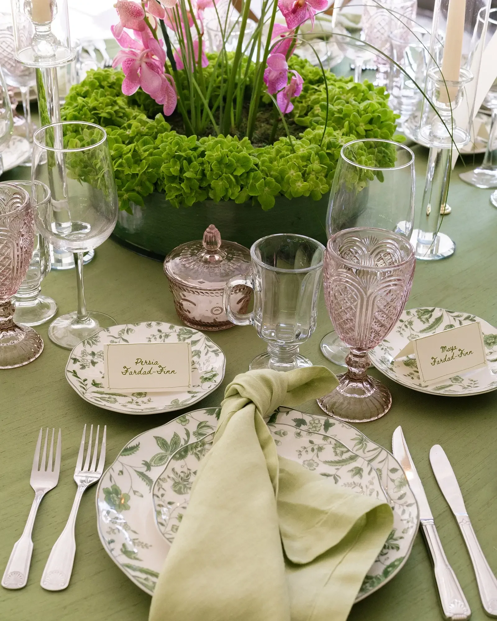 Elegant table setting with floral patterned plates, a pale green napkin, crystal glasses, and a centerpiece of pink flowers and green foliage on a green tablecloth.