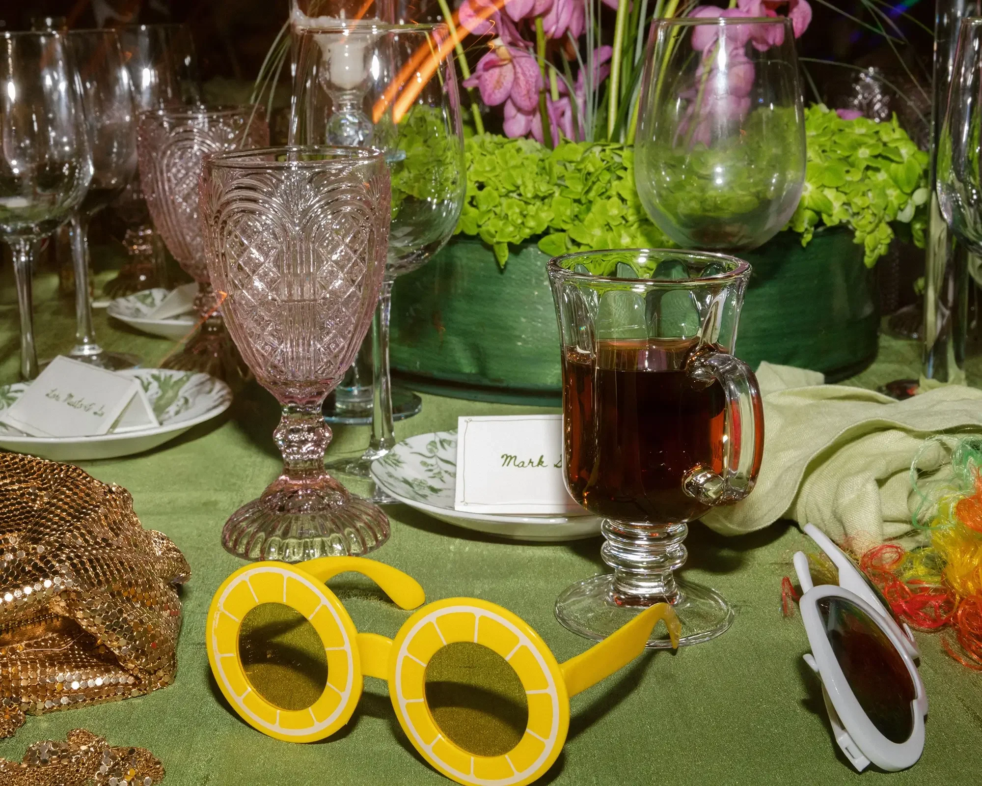 A decorative wedding table setting with pink and green glassware, a floral centerpiece, a cup of dcoffee, a pair of yellow novelty sunglasses, and other festive accessories.