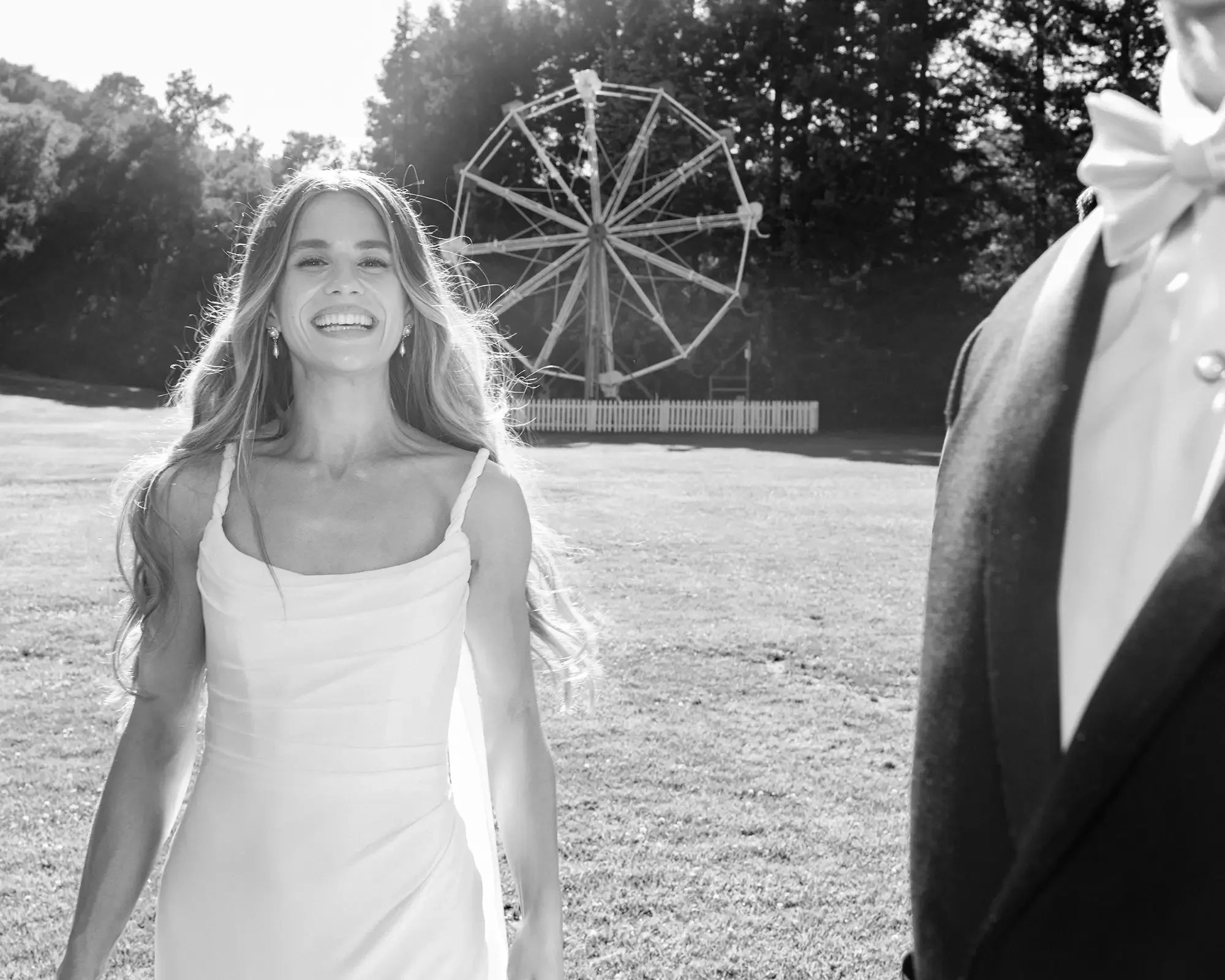 A woman in a wedding dress smiling outdoors, with a ferris wheel in the background and a groom in a tuxedo partially visible.