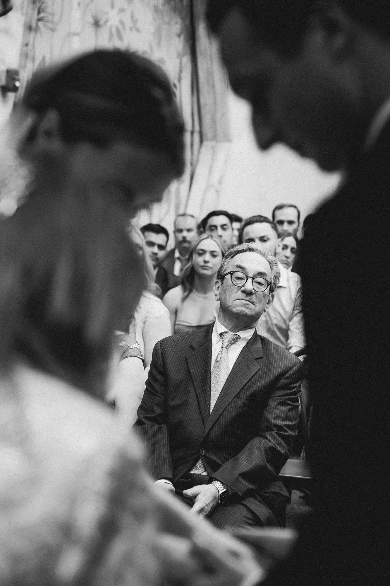 Black and white photo showing the father of the bride in a suit with glasses sitting during the wedding ceremony filled with people. The man appears to be listening or contemplating, with a serious or annoyed expression. 