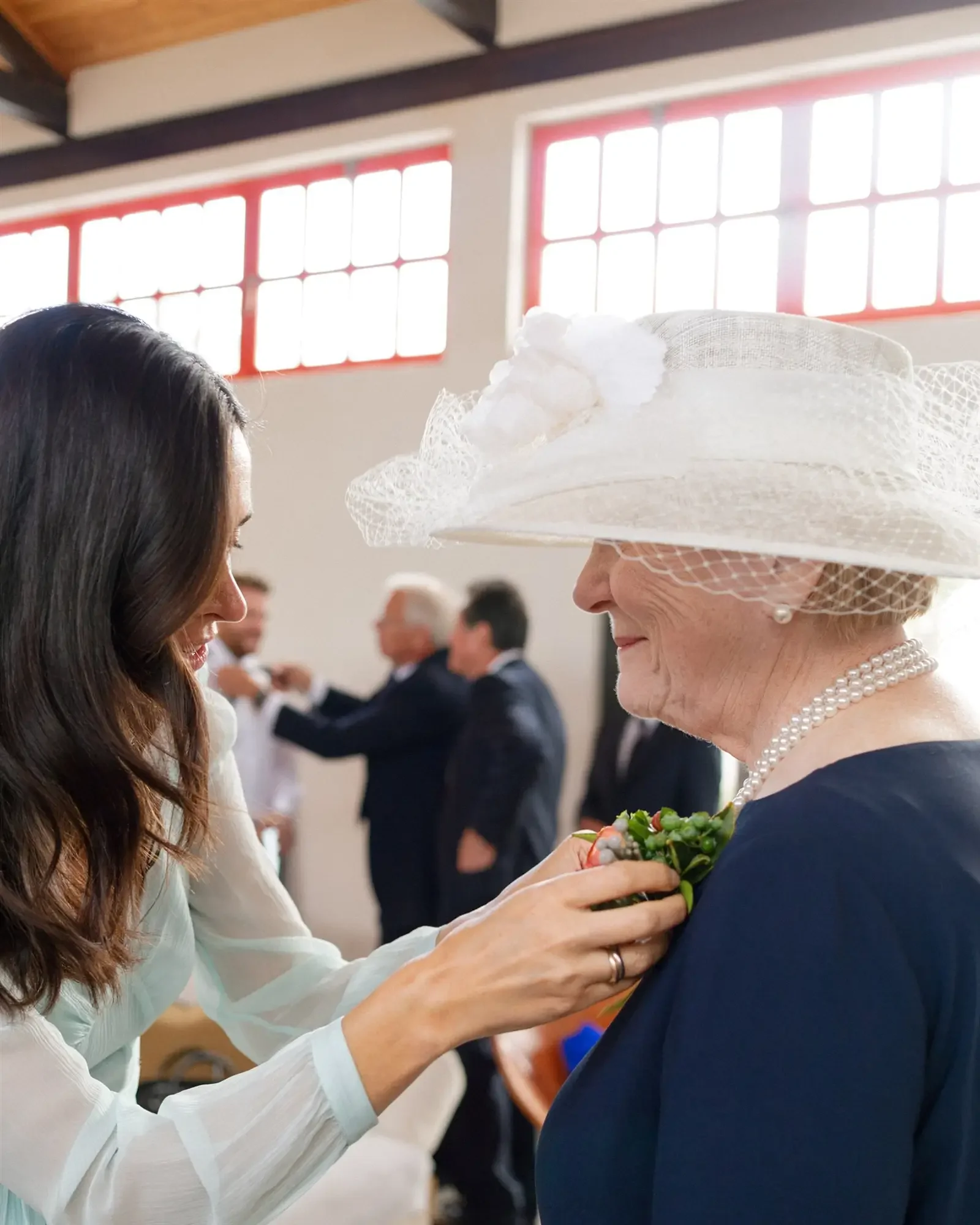 A young woman pinning a corsage on an elderly woman wearing a large white hat at a social gathering.